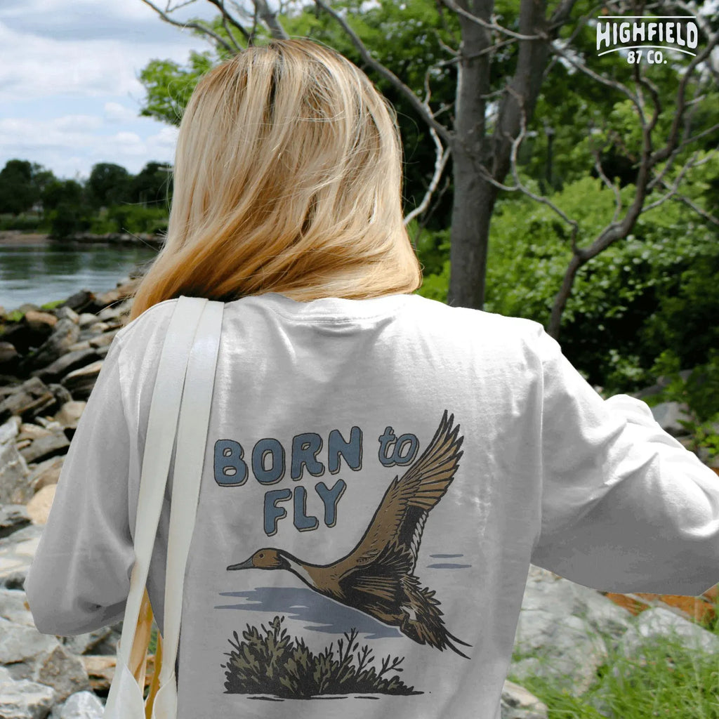 Brown pintail duck with detailed wings flying over wetland brush. Text above reads "Born to Fly" in blue retro font. For hunting or nature gear design.