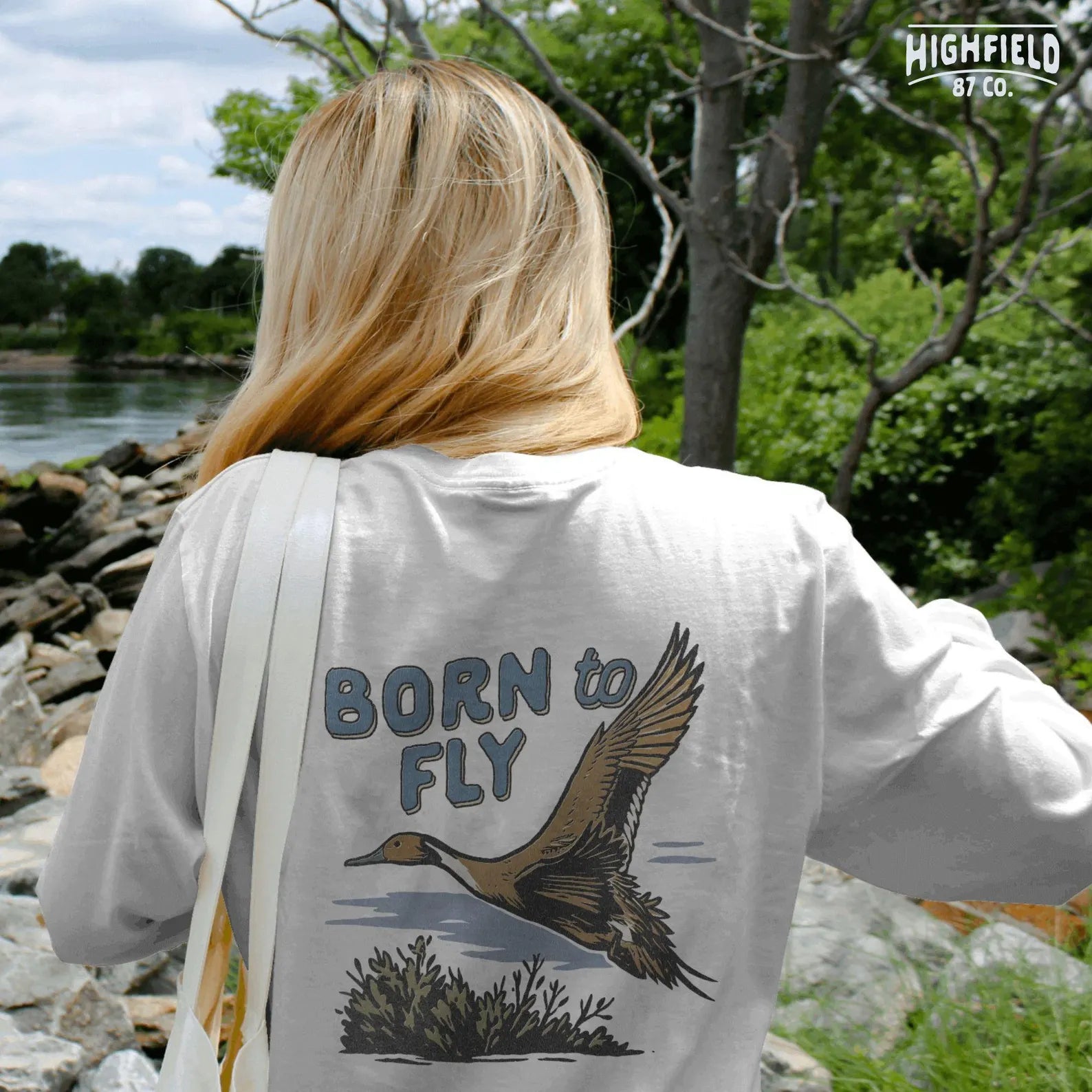 Brown pintail duck with detailed wings flying over wetland brush. Text above reads "Born to Fly" in blue retro font. For hunting or nature gear design.