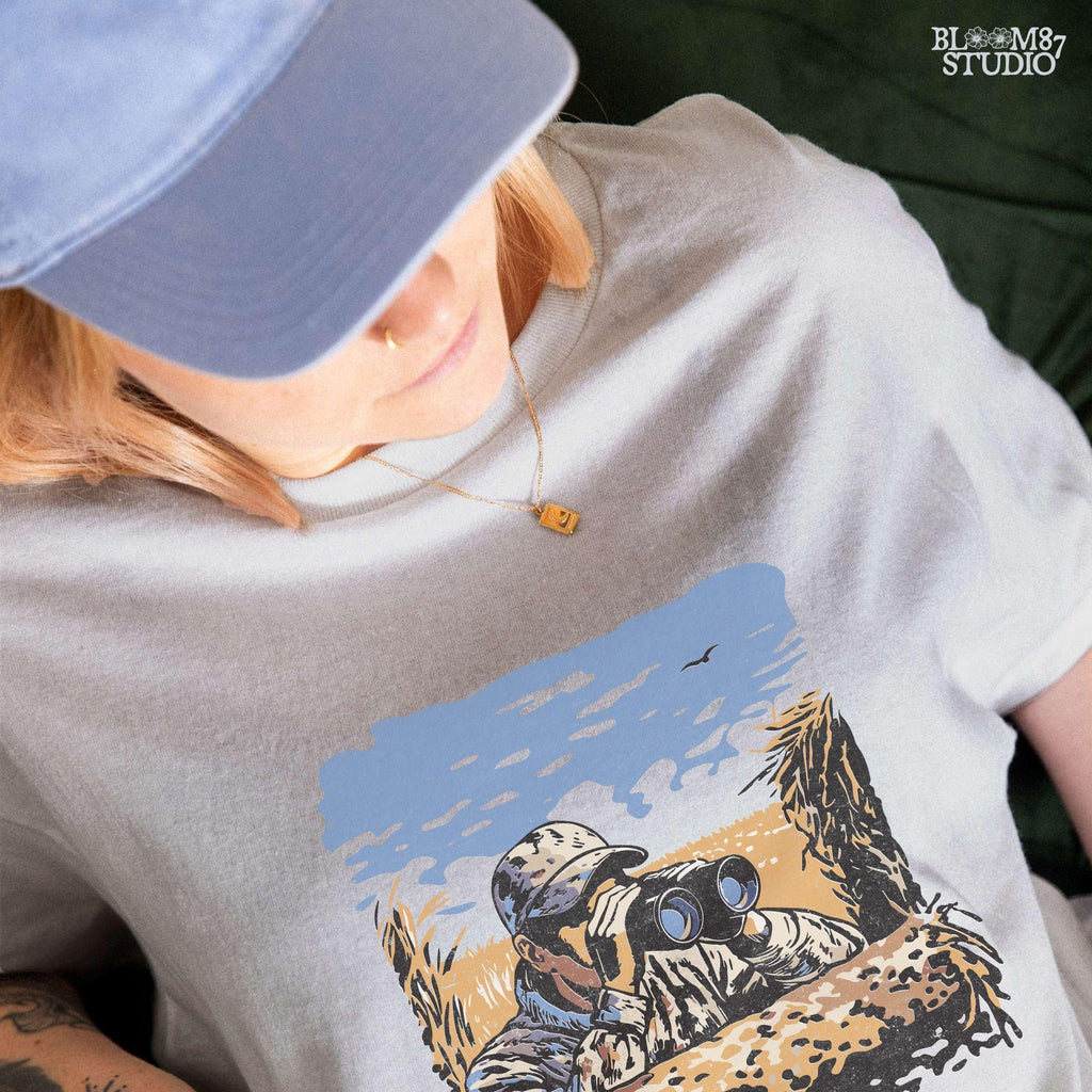 “Illustrated camouflage hunter peeking through binoculars from a duck blind, surrounded by tall grass, with a blue sky and golden field background. A detailed digital hunting-themed design, perfect for outdoorsmen, wildlife lovers, and hunting gear.”