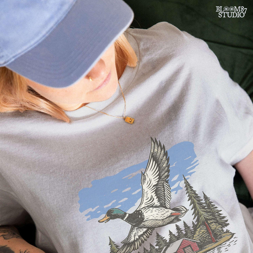 Vintage illustration of a mallard duck flying over a red cabin by the lake, surrounded by pine trees. Perfect for rustic lodge decor and wildlife art prints.