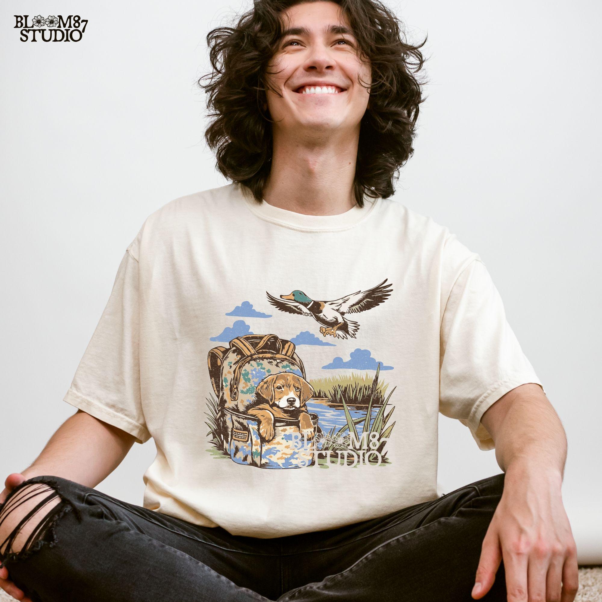 Illustration of a hunting dog resting in a camouflage bag near a pond, with a mallard duck flying overhead in a peaceful outdoor setting.