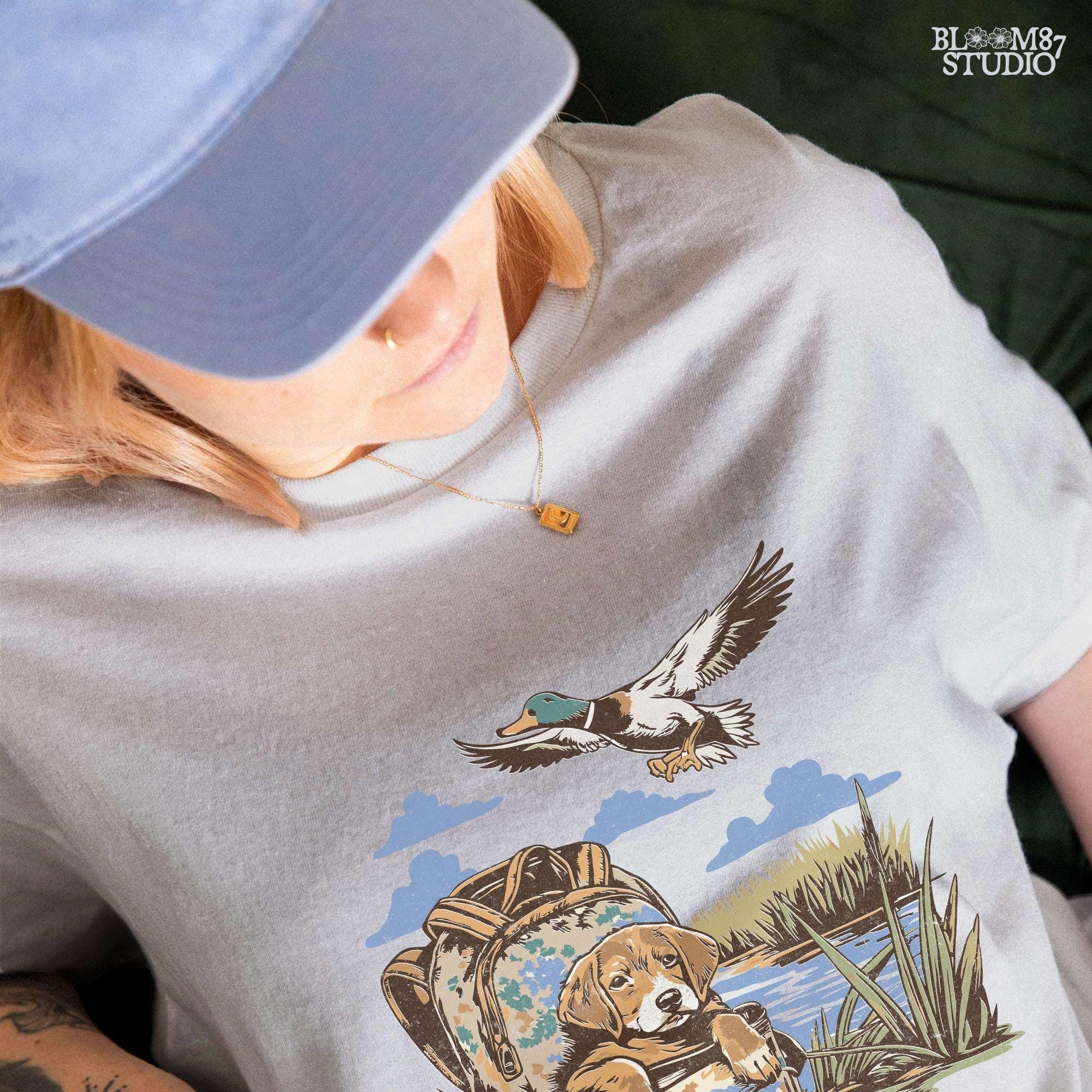 Illustration of a hunting dog resting in a camouflage bag near a pond, with a mallard duck flying overhead in a peaceful outdoor setting.