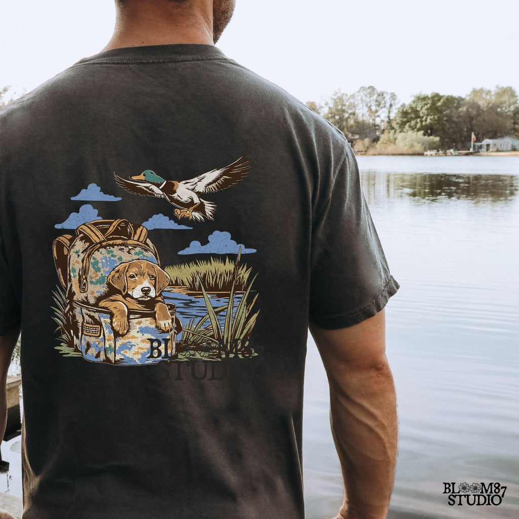 Illustration of a hunting dog resting in a camouflage bag near a pond, with a mallard duck flying overhead in a peaceful outdoor setting.