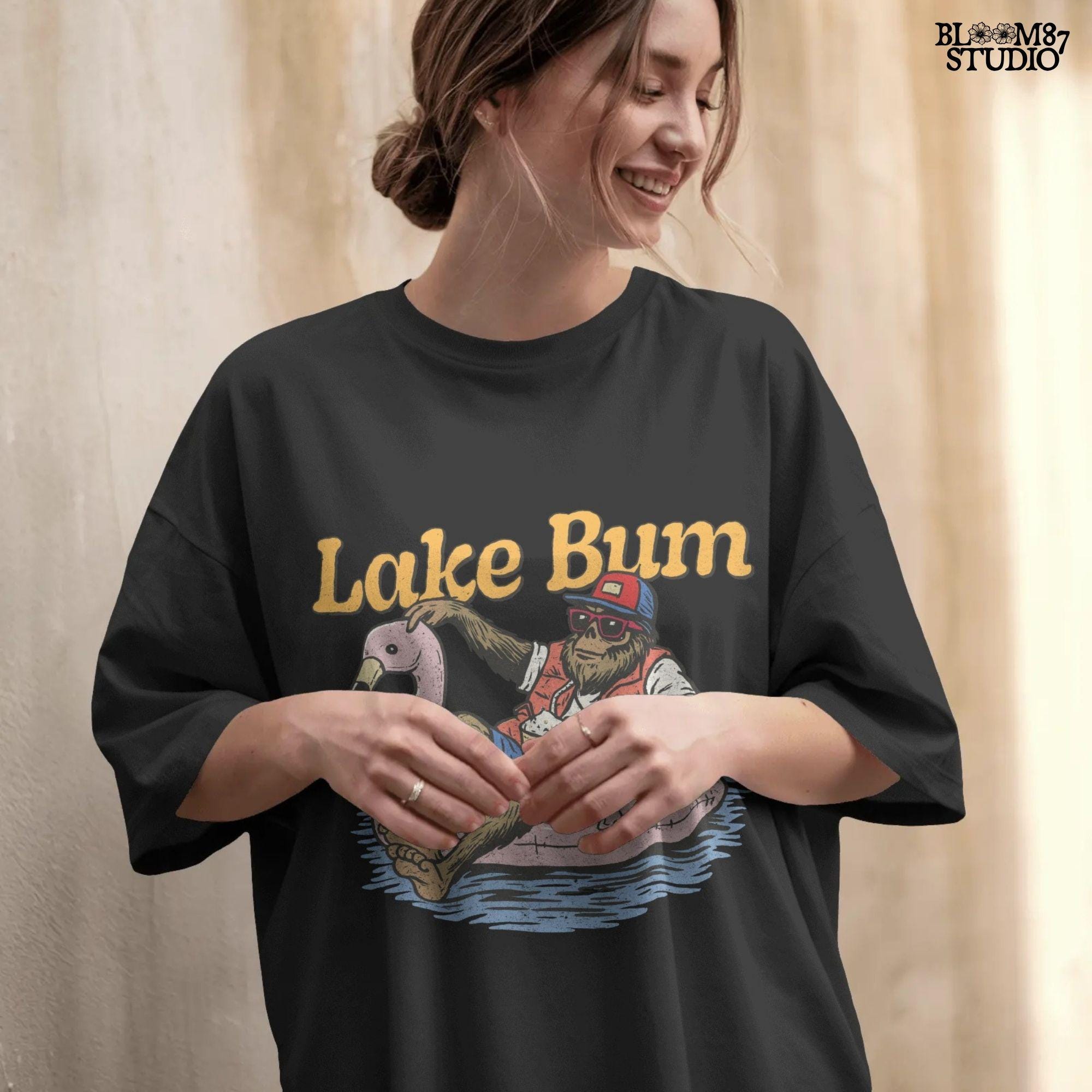 Bigfoot lounging on a pink flamingo float with a beer, wearing summer gear and sunglasses, with “Lake Bum” text above.