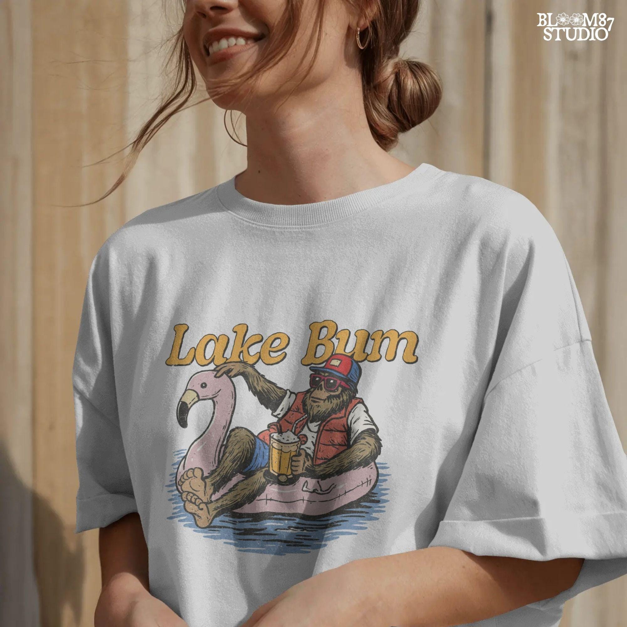 Bigfoot lounging on a pink flamingo float with a beer, wearing summer gear and sunglasses, with “Lake Bum” text above.