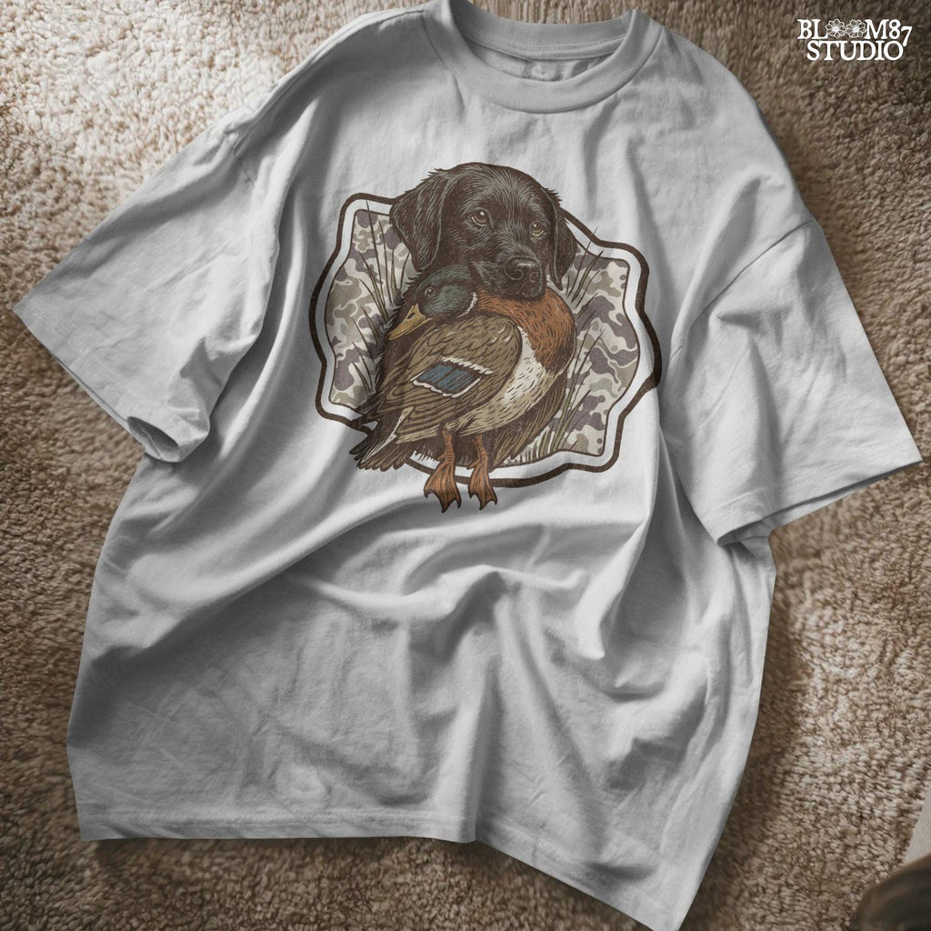 Black Labrador retriever holding a mallard duck with a camouflage background, perfect for hunting or outdoors-themed designs.