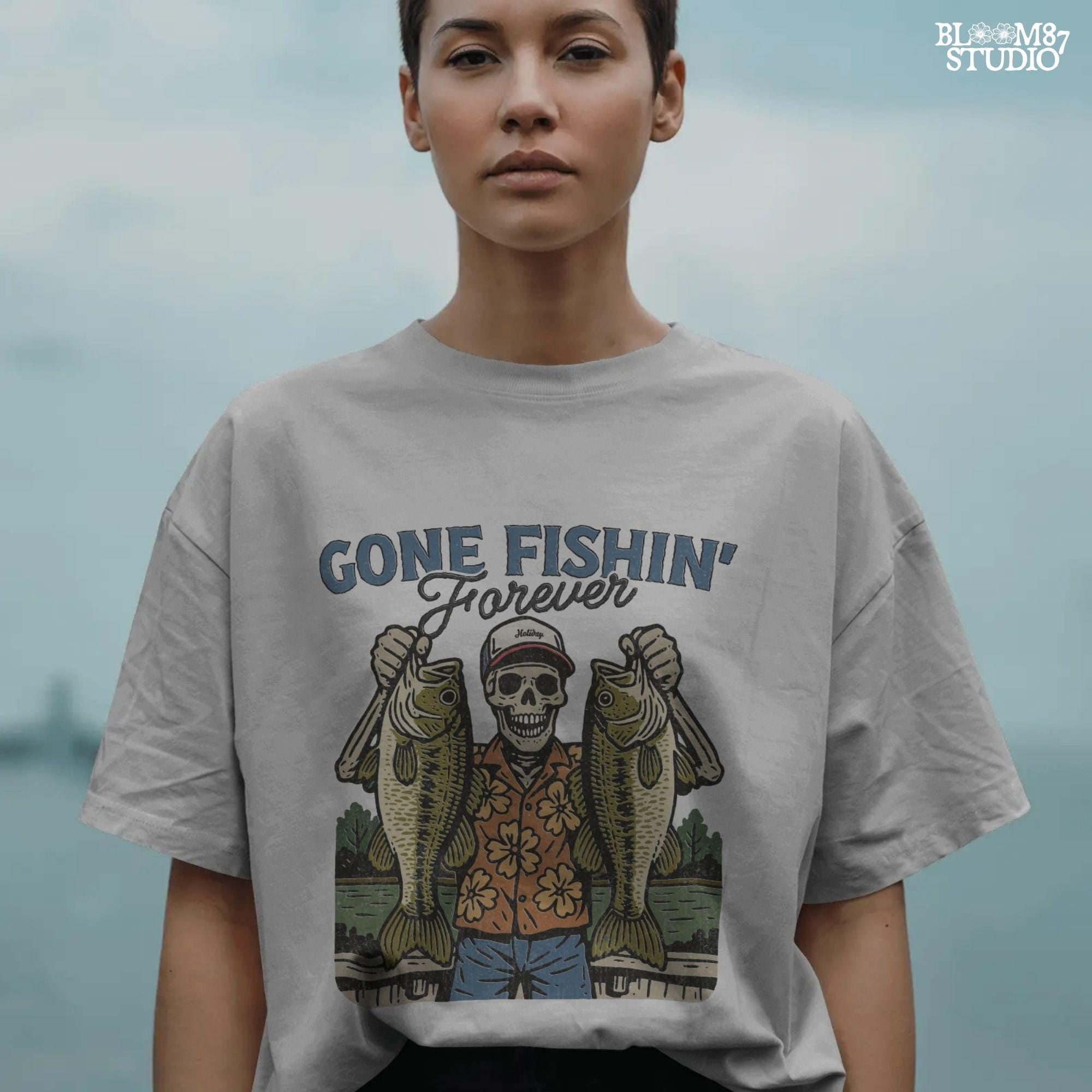 A skeleton wearing a red cap and orange floral shirt stands on a dock holding two large bass fish with the words “Gone Fishin’ Forever” above.