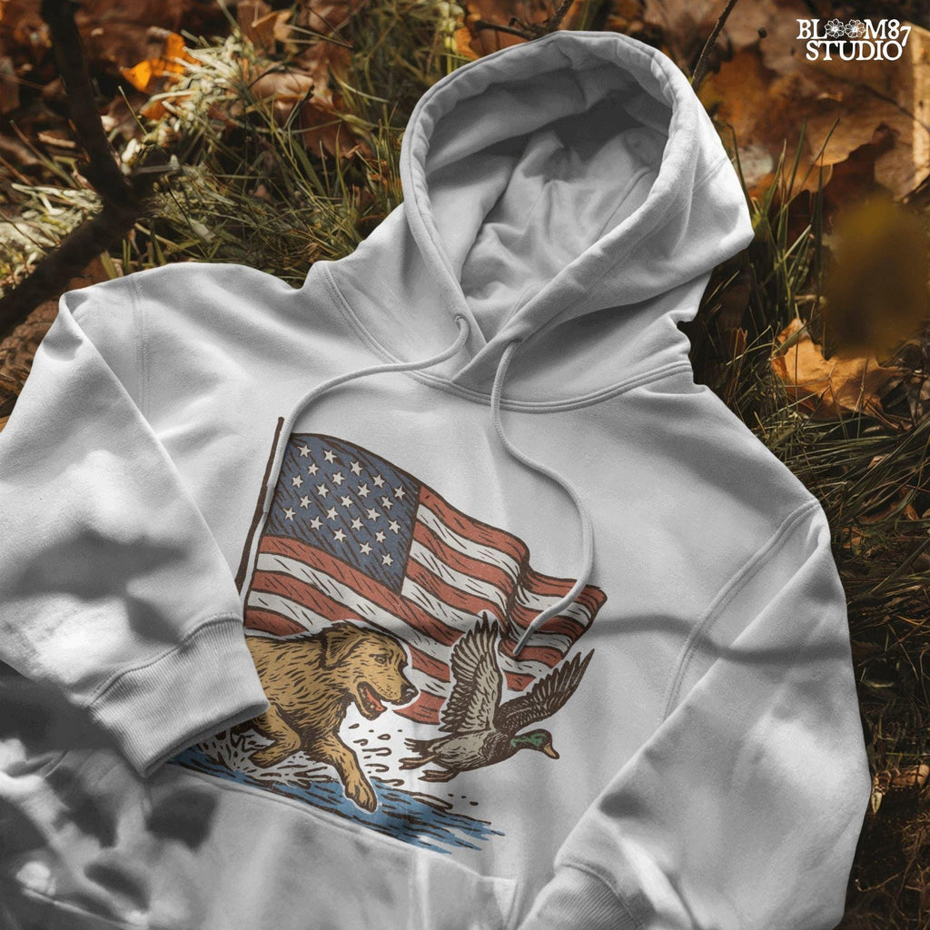 A yellow Labrador retriever splashing in water next to a flying duck, with a waving American flag in the background.