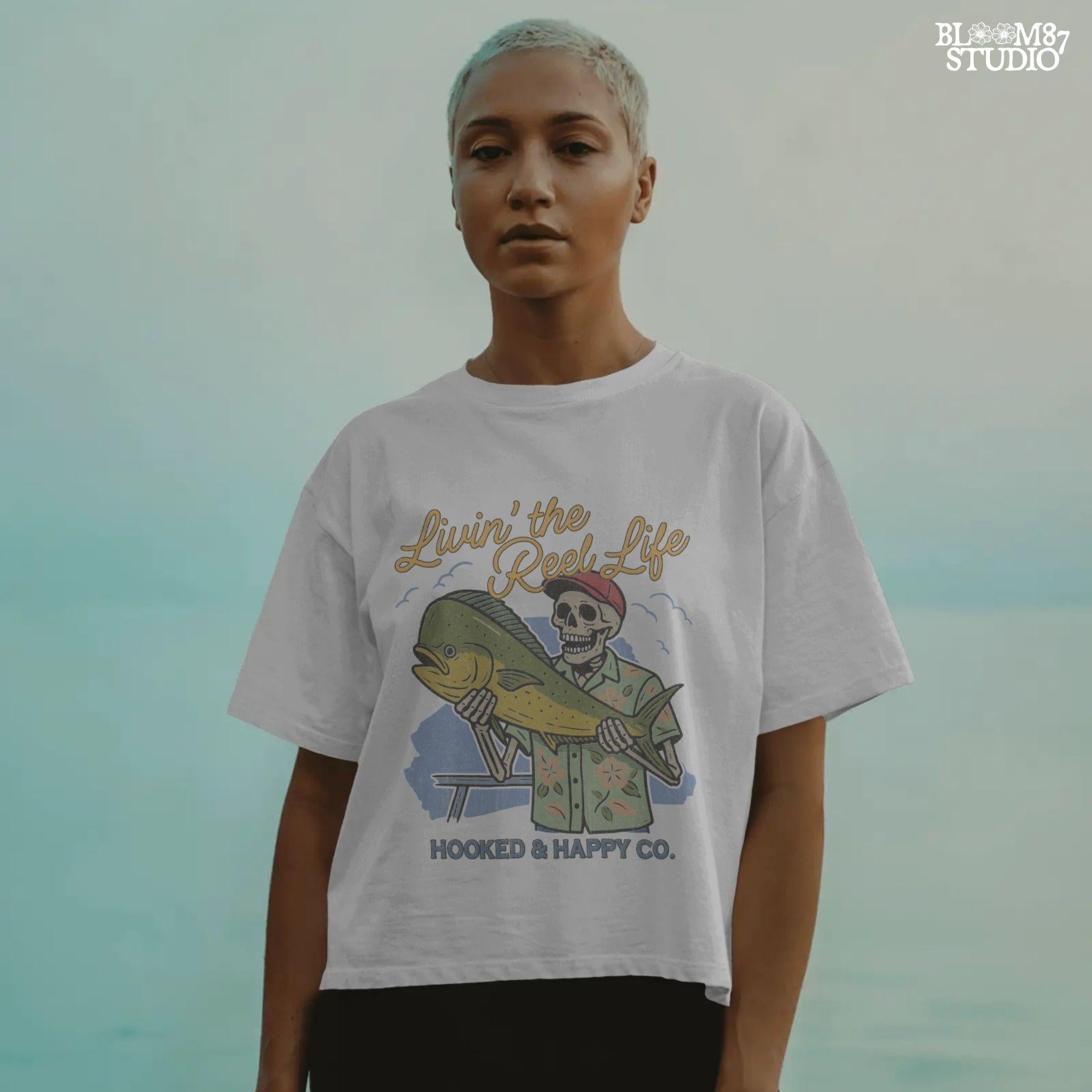 Cartoon skeleton in tropical shirt holding a mahi-mahi fish with text “Livin’ the Reel Life” above and “Hooked & Happy Co.” below.