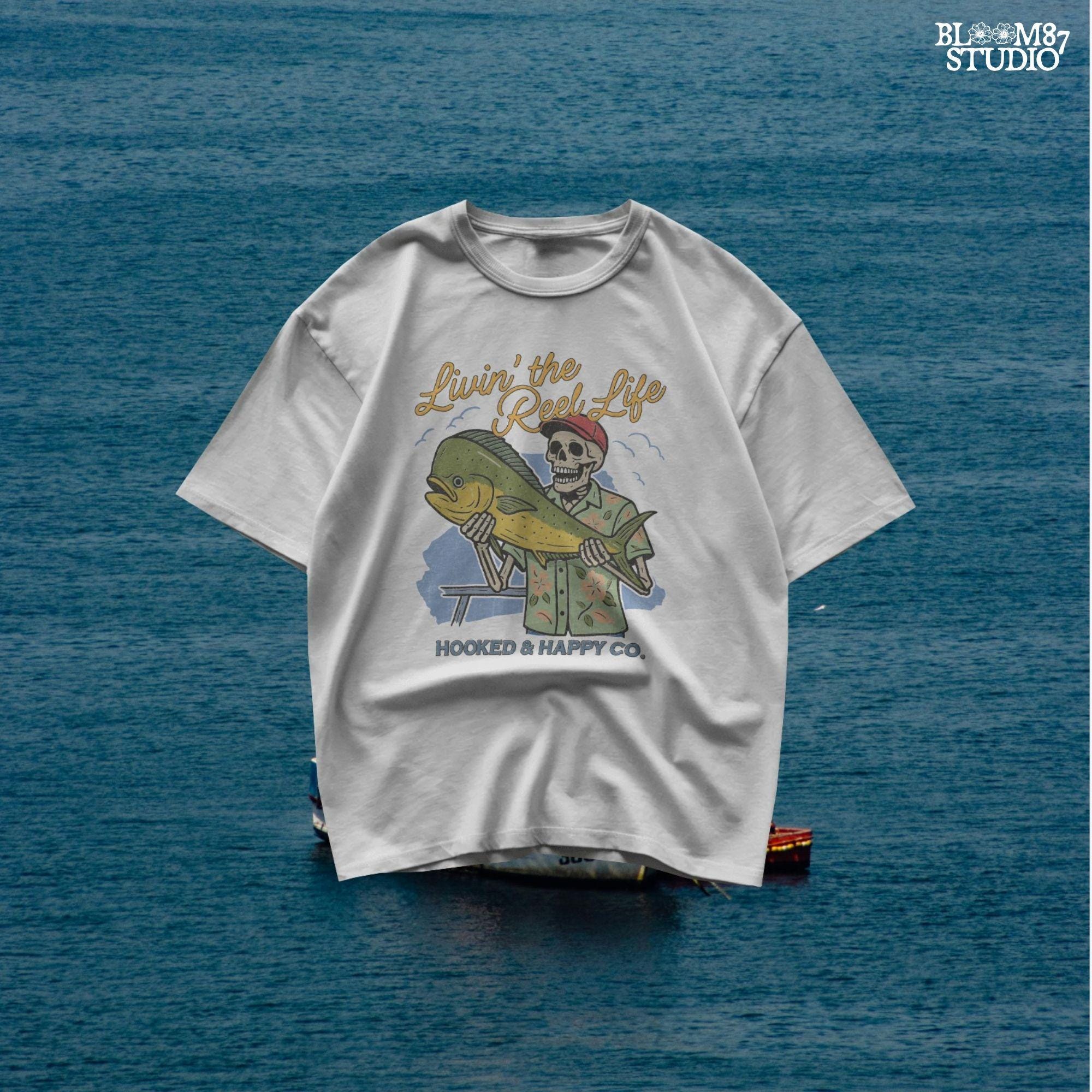 Cartoon skeleton in tropical shirt holding a mahi-mahi fish with text “Livin’ the Reel Life” above and “Hooked & Happy Co.” below.