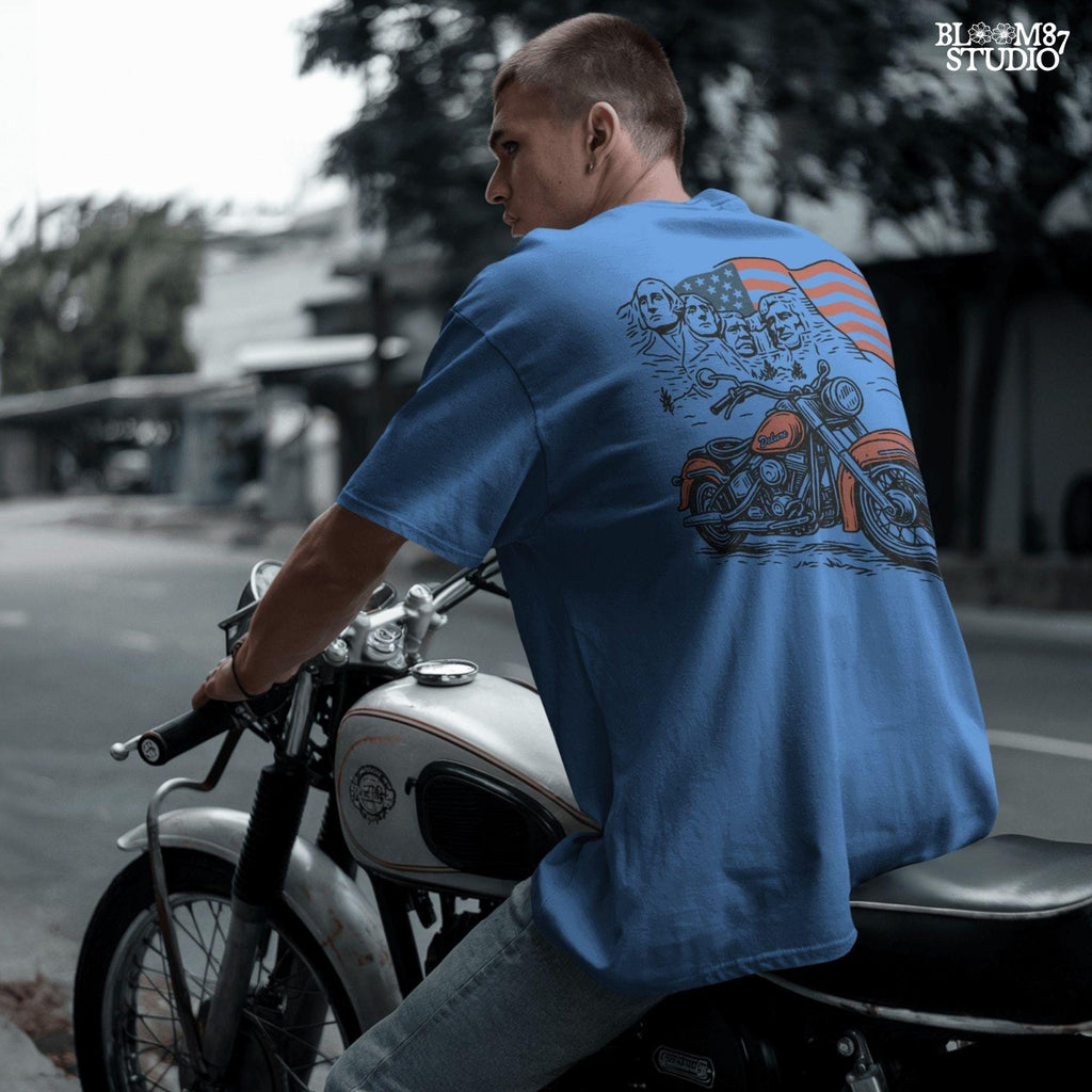 Vintage motorcycle graphic in front of Mount Rushmore and a waving American flag, styled in patriotic colors with retro biker design elements.