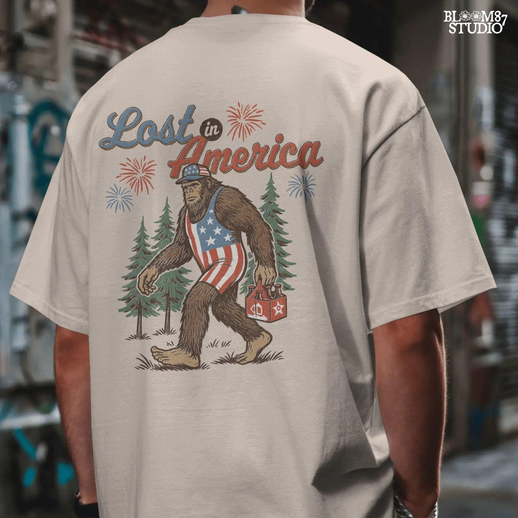 Bigfoot wearing an American flag outfit, carrying a six-pack of beer, walking through pine trees with fireworks in the sky and the phrase “Lost in America.”