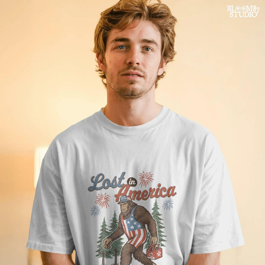 Bigfoot wearing an American flag outfit, carrying a six-pack of beer, walking through pine trees with fireworks in the sky and the phrase “Lost in America.”