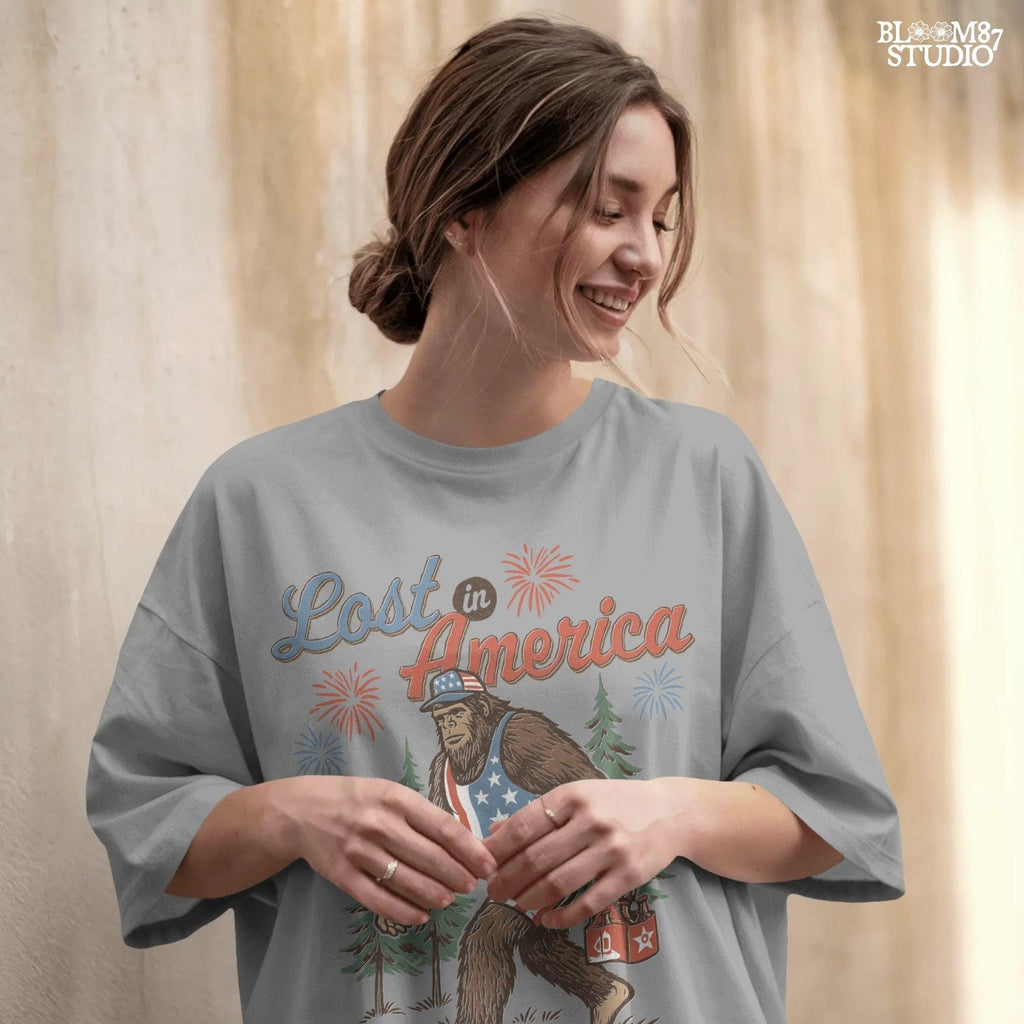 Bigfoot wearing an American flag outfit, carrying a six-pack of beer, walking through pine trees with fireworks in the sky and the phrase “Lost in America.”