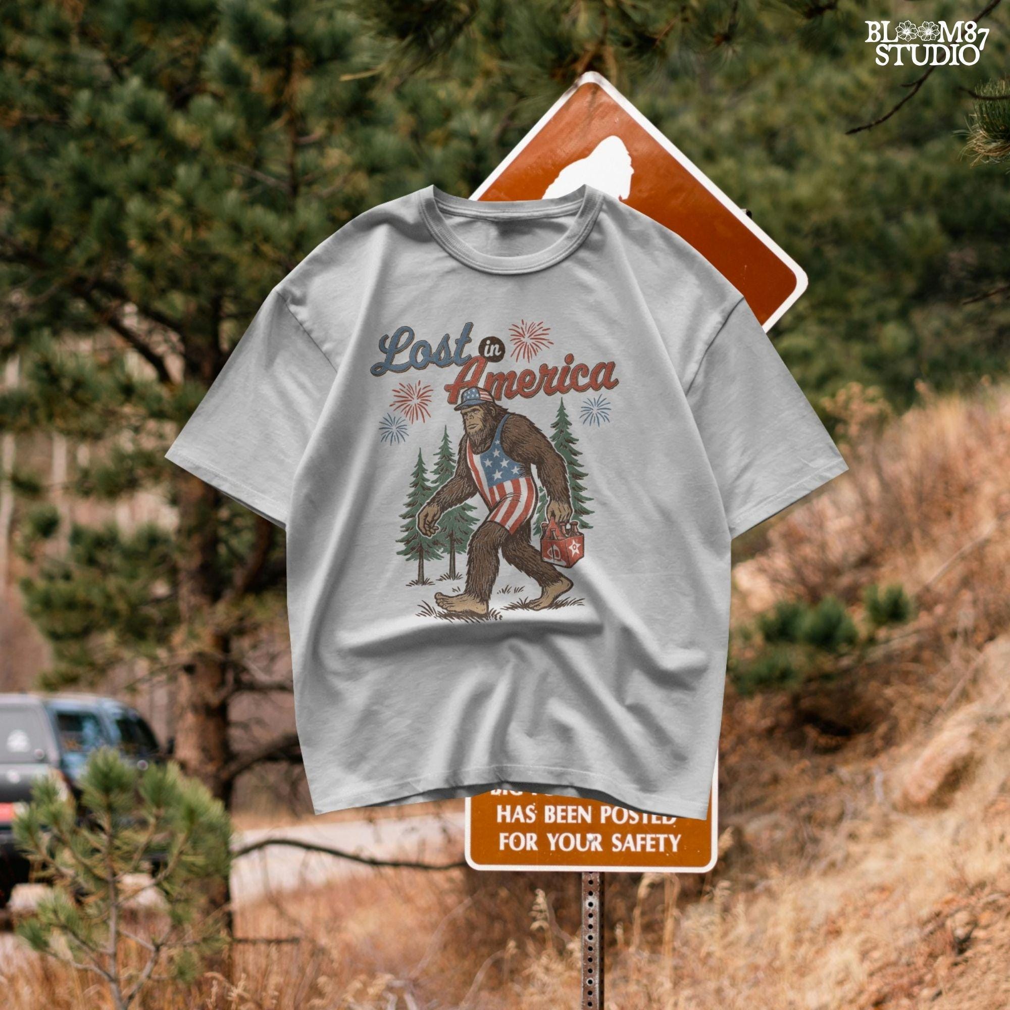Bigfoot wearing an American flag outfit, carrying a six-pack of beer, walking through pine trees with fireworks in the sky and the phrase “Lost in America.”