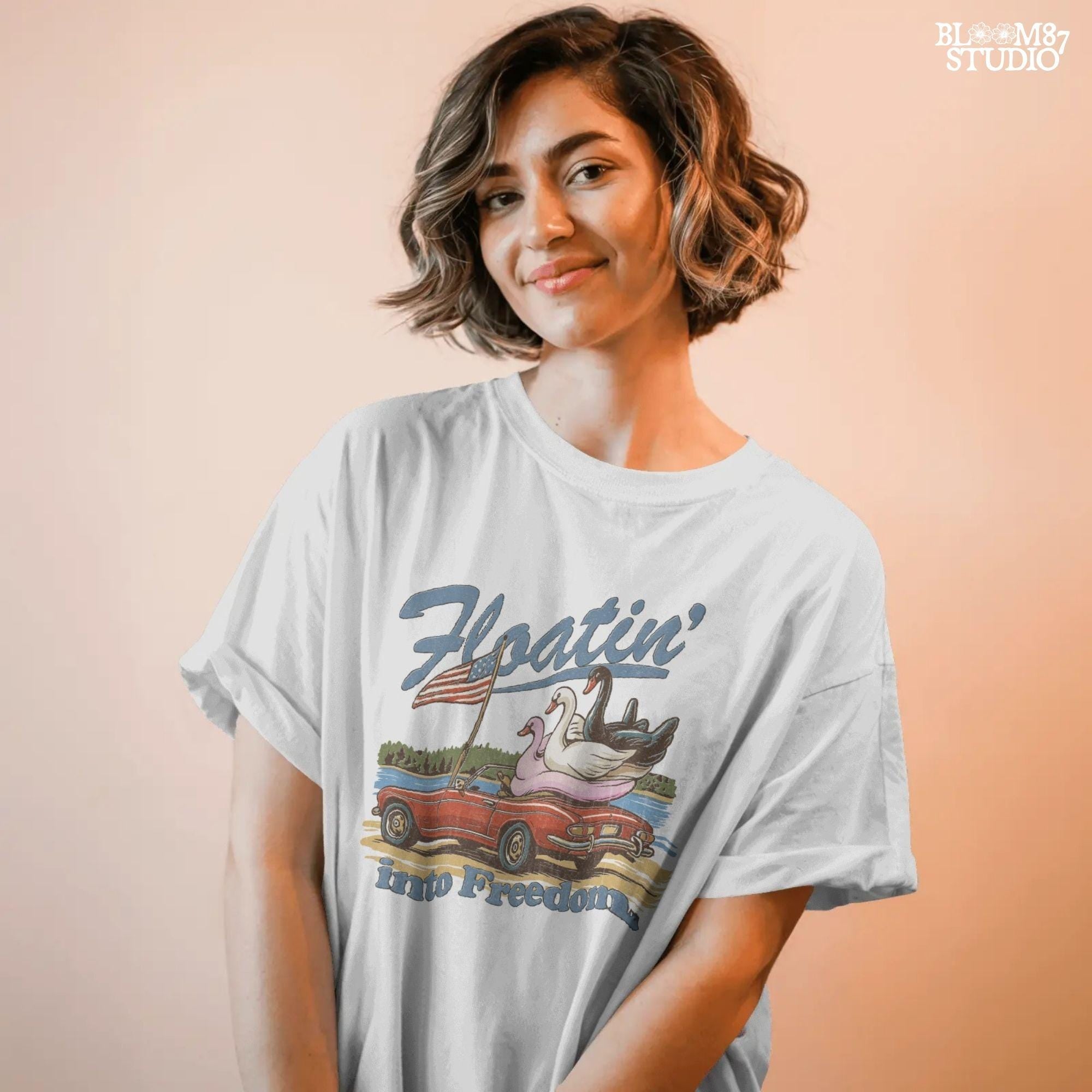 A red convertible car on the beach carrying three swan pool floats with an American flag, with the text “Floatin’ into Freedom” in a playful patriotic theme.