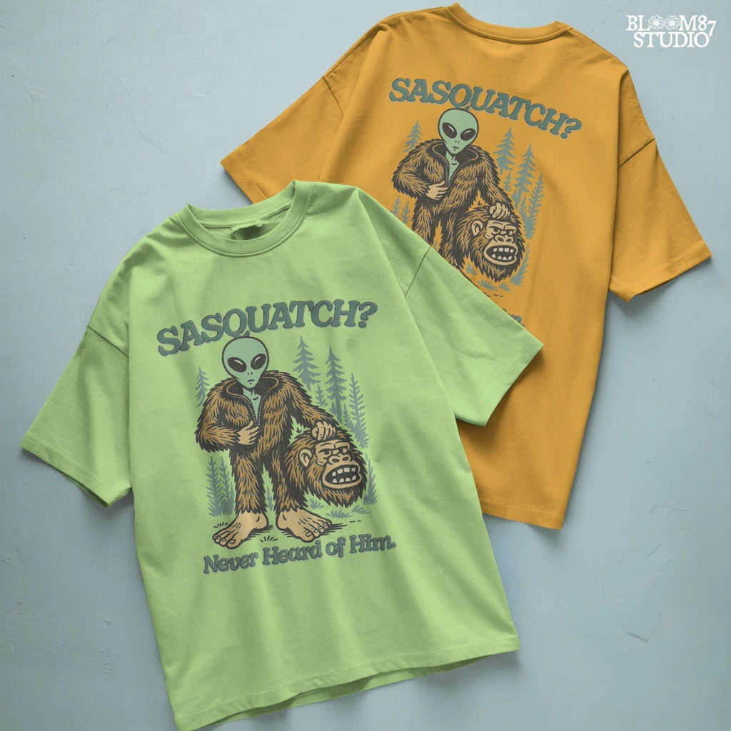 Funny alien unzipping a Bigfoot costume with Sasquatch head in hand, forest trees in background, and text “Sasquatch? Never Heard of Him.”