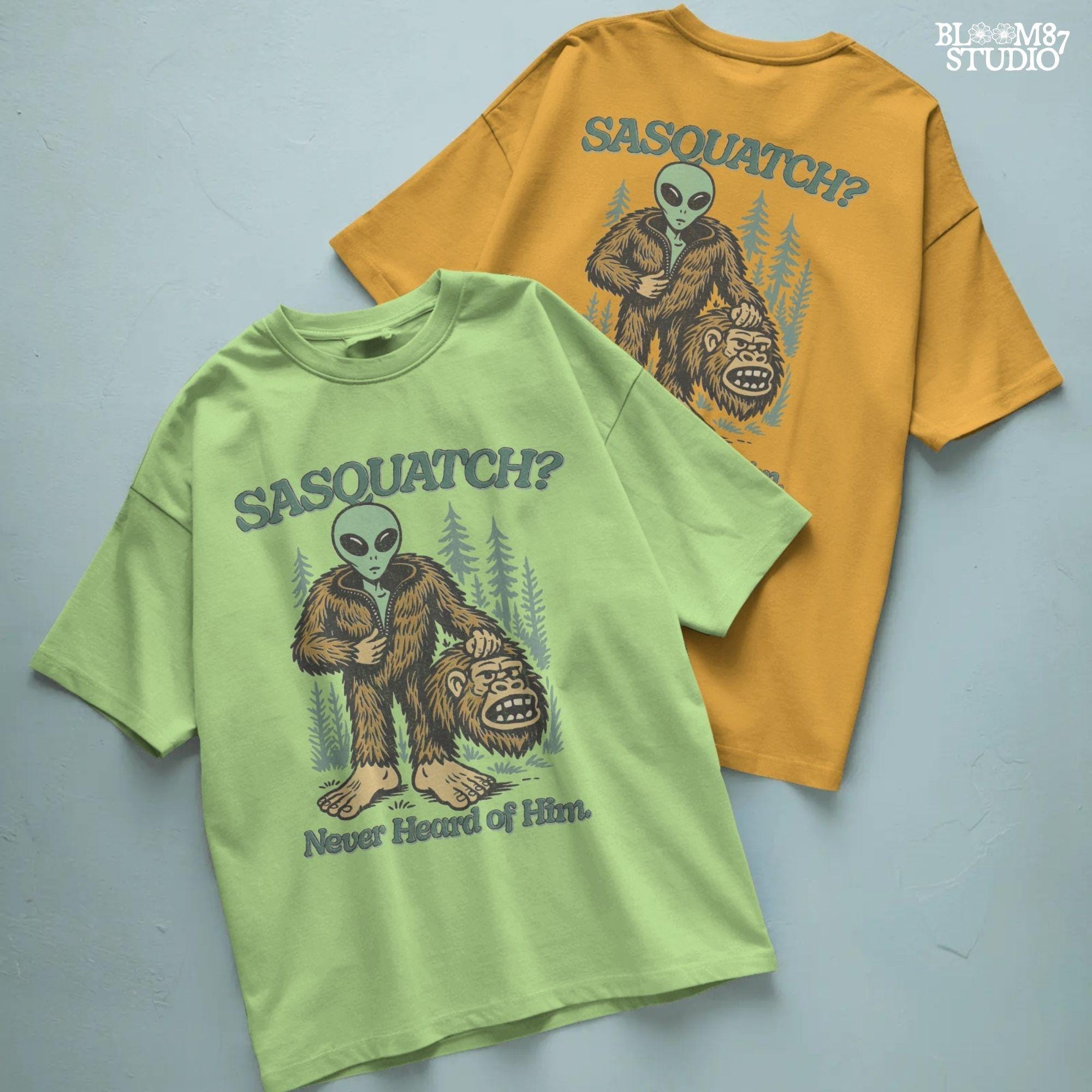 Funny alien unzipping a Bigfoot costume with Sasquatch head in hand, forest trees in background, and text “Sasquatch? Never Heard of Him.”