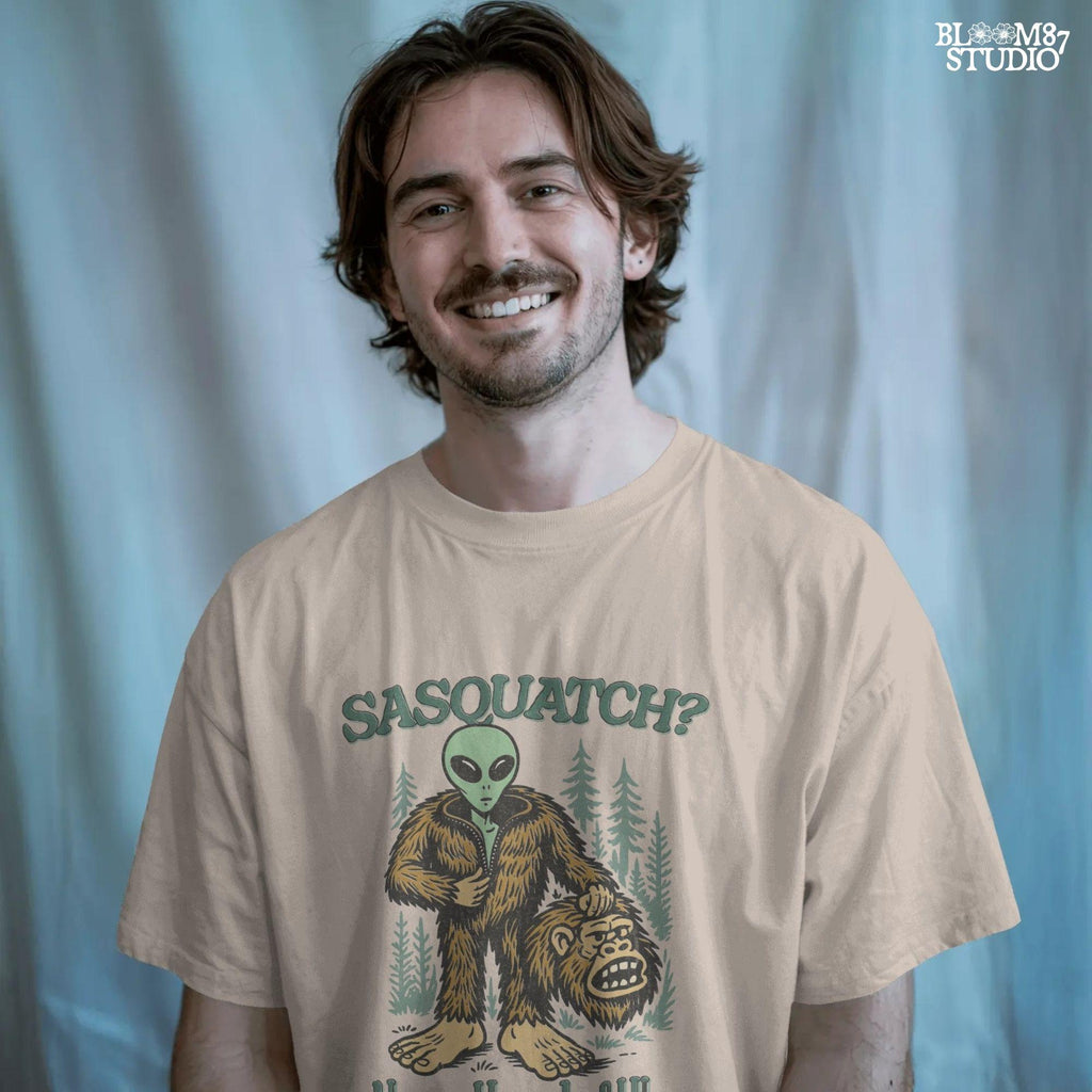 Funny alien unzipping a Bigfoot costume with Sasquatch head in hand, forest trees in background, and text “Sasquatch? Never Heard of Him.”