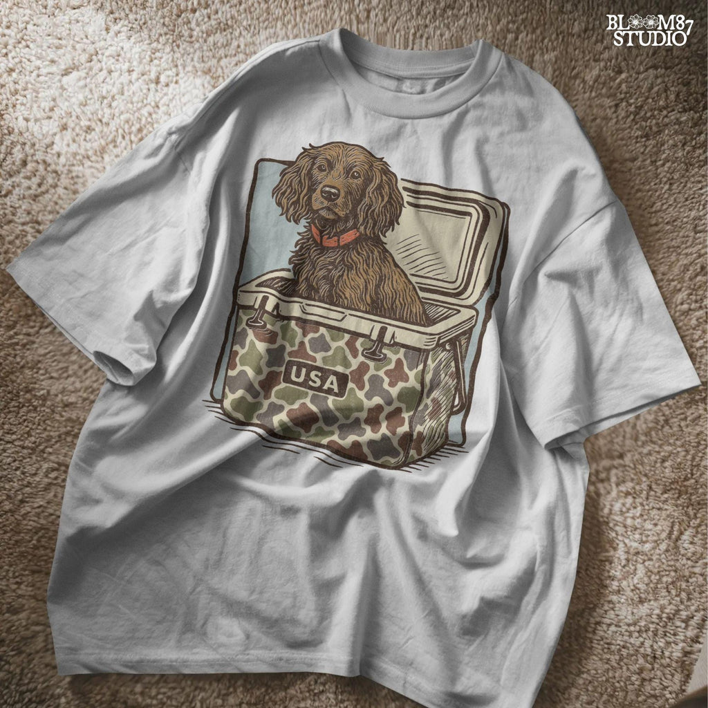 Hunting dog with curly fur sitting inside a camouflage cooler marked “USA” in a retro-style outdoors graphic.