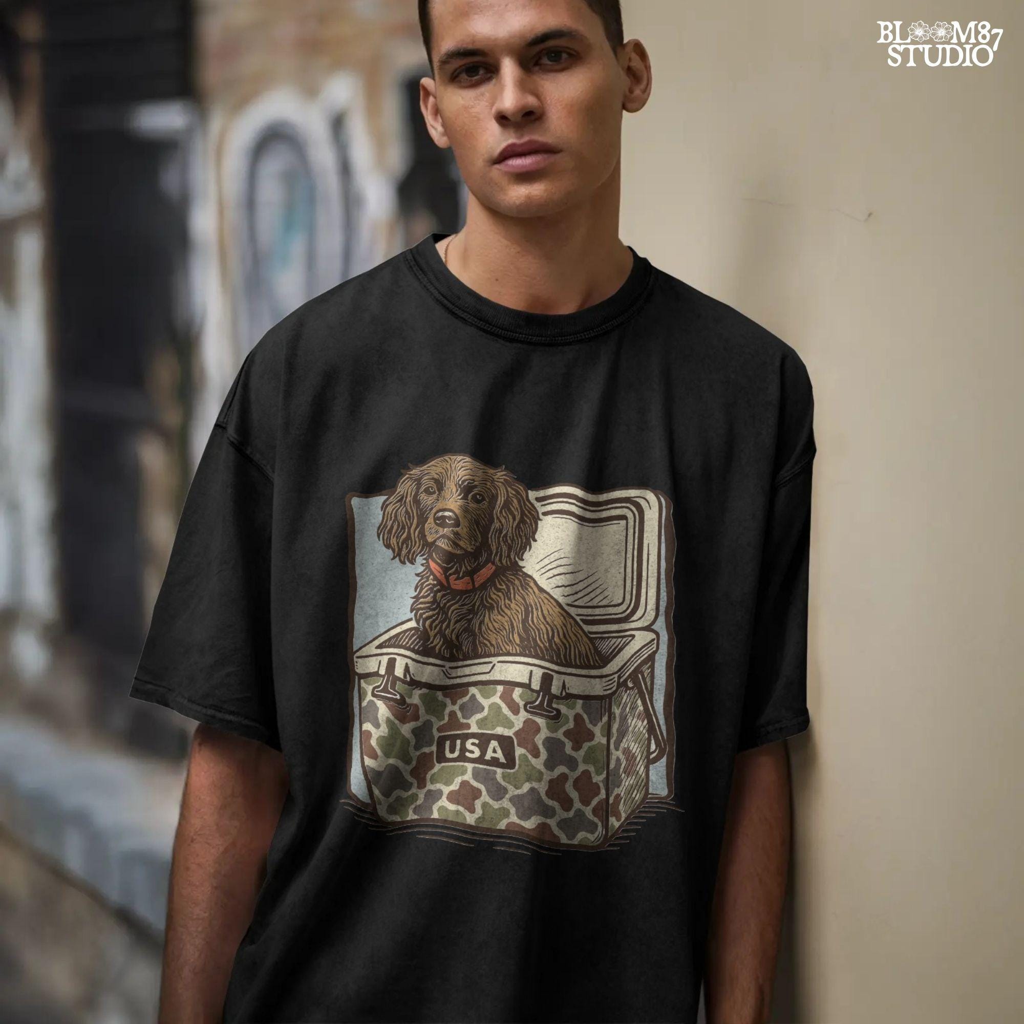 Hunting dog with curly fur sitting inside a camouflage cooler marked “USA” in a retro-style outdoors graphic.