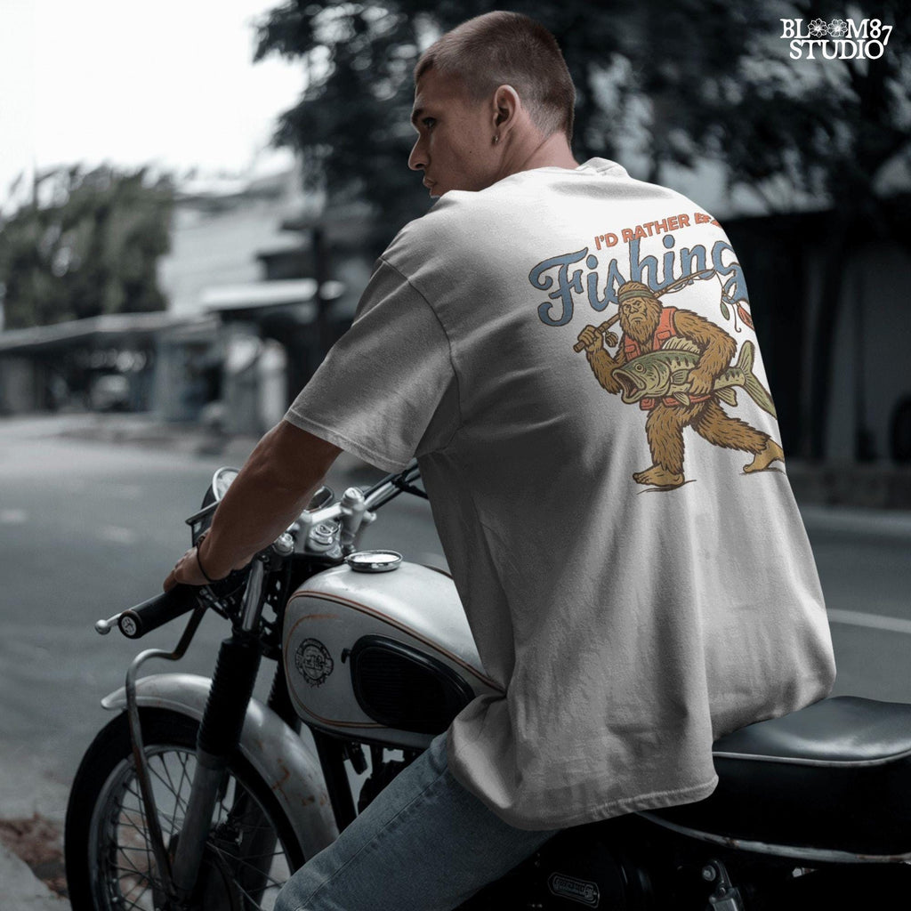 Funny vintage illustration of Bigfoot wearing a fishing vest, holding a giant bass and fishing rod, with text that reads “I’d Rather Be Fishing.”