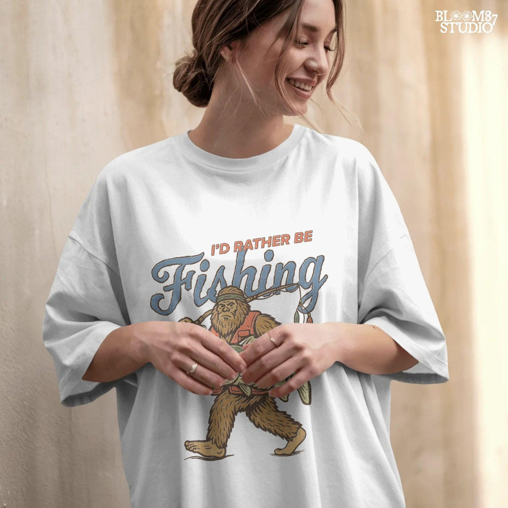 Funny vintage illustration of Bigfoot wearing a fishing vest, holding a giant bass and fishing rod, with text that reads “I’d Rather Be Fishing.”