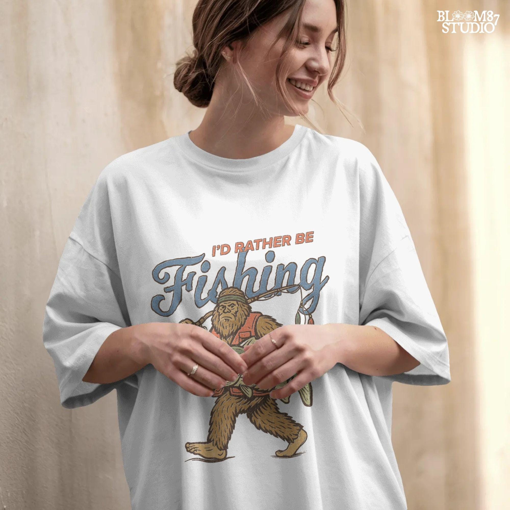 Funny vintage illustration of Bigfoot wearing a fishing vest, holding a giant bass and fishing rod, with text that reads “I’d Rather Be Fishing.”