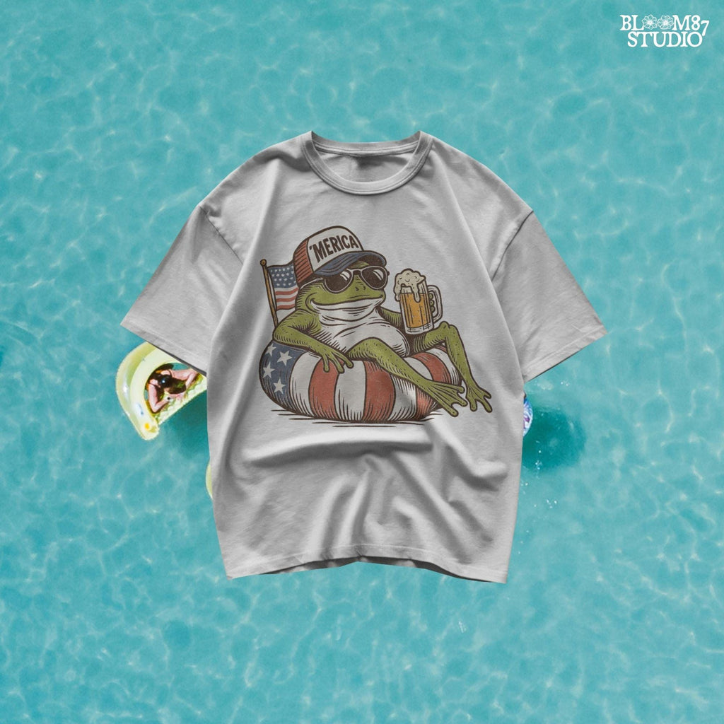 A vintage-style cartoon frog lounging on a patriotic inner tube, wearing a ‘Merica cap and sunglasses, holding a mug of beer with an American flag in the background.
