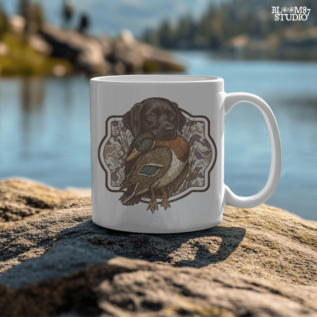 Black Labrador retriever holding a mallard duck with a camouflage background, perfect for hunting or outdoors-themed designs.