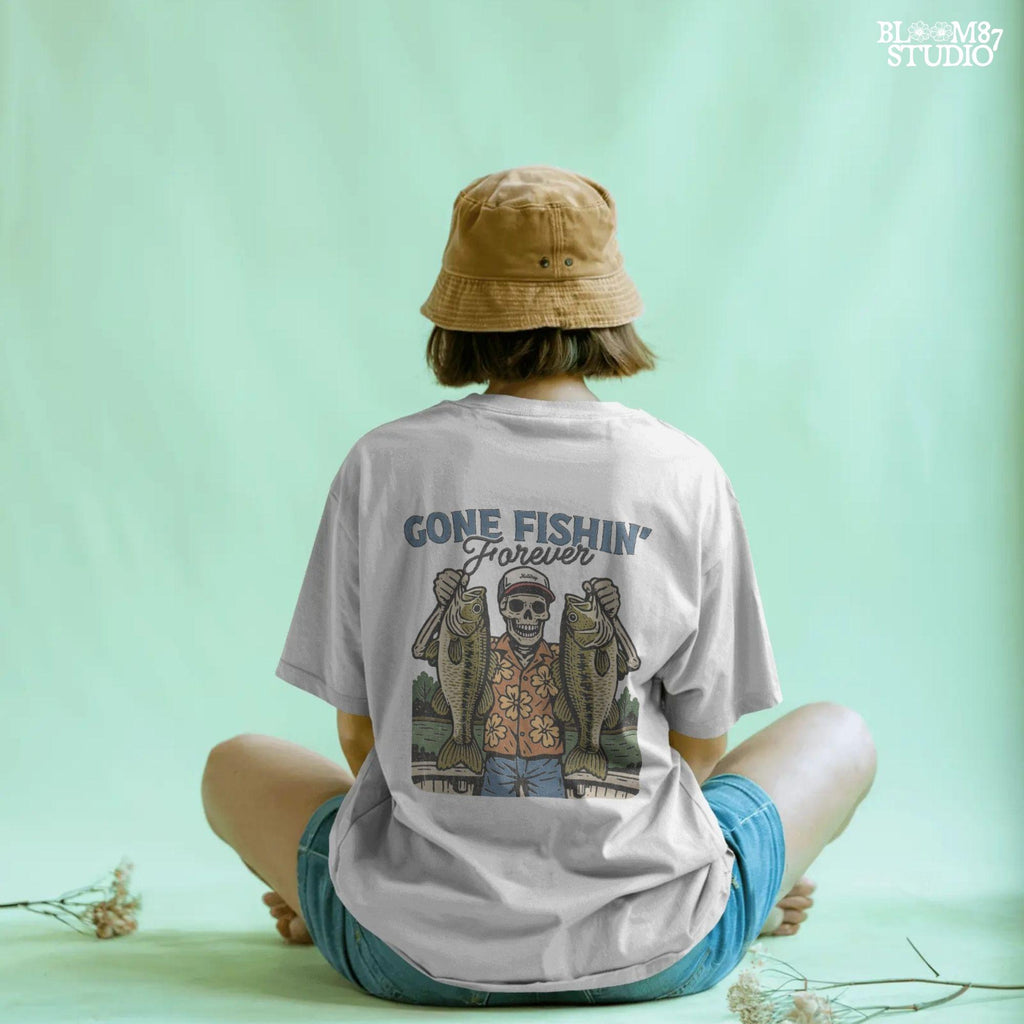 A skeleton wearing a red cap and orange floral shirt stands on a dock holding two large bass fish with the words “Gone Fishin’ Forever” above.