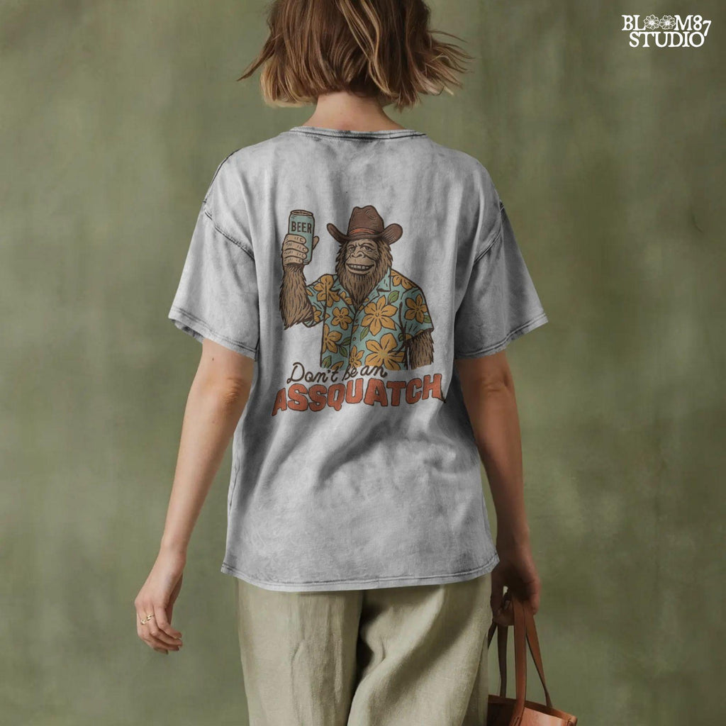 Retro cartoon Sasquatch in floral shirt holding a beer can with text “Don’t Be an Assquatch,” perfect for funny camping or summer designs