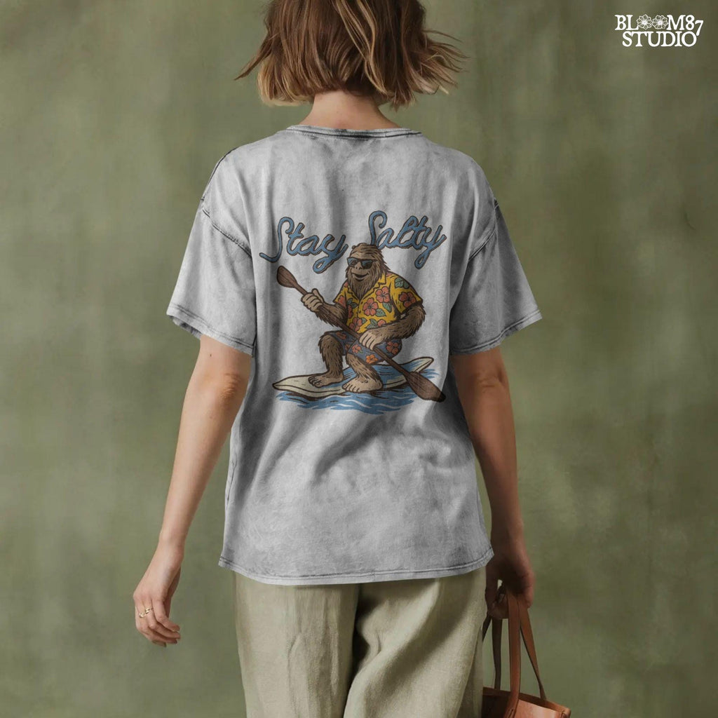 Cartoon Bigfoot wearing a Hawaiian shirt and sunglasses, paddleboarding on a surfboard with text “Stay Salty” – retro beach design PNG for sublimation.