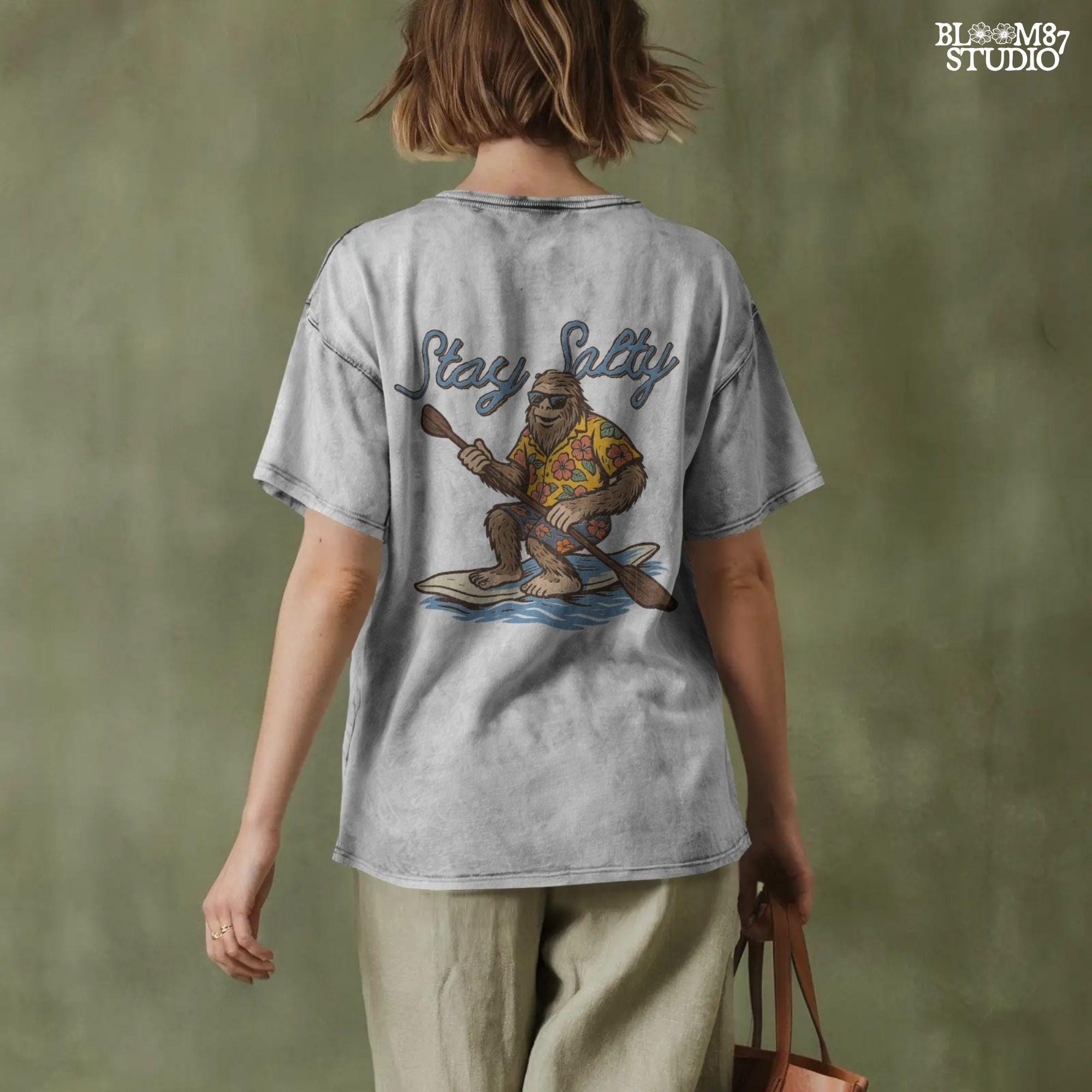 Cartoon Bigfoot wearing a Hawaiian shirt and sunglasses, paddleboarding on a surfboard with text “Stay Salty” – retro beach design PNG for sublimation.