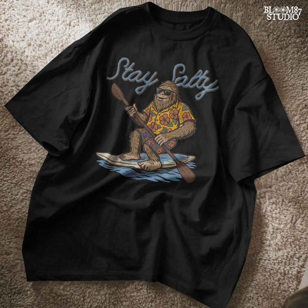 Cartoon Bigfoot wearing a Hawaiian shirt and sunglasses, paddleboarding on a surfboard with text “Stay Salty” – retro beach design PNG for sublimation.