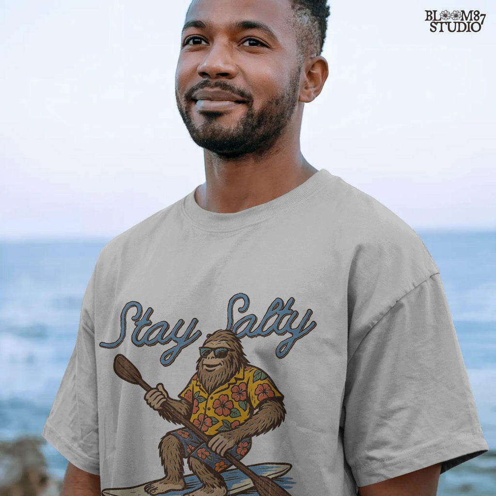 Cartoon Bigfoot wearing a Hawaiian shirt and sunglasses, paddleboarding on a surfboard with text “Stay Salty” – retro beach design PNG for sublimation.