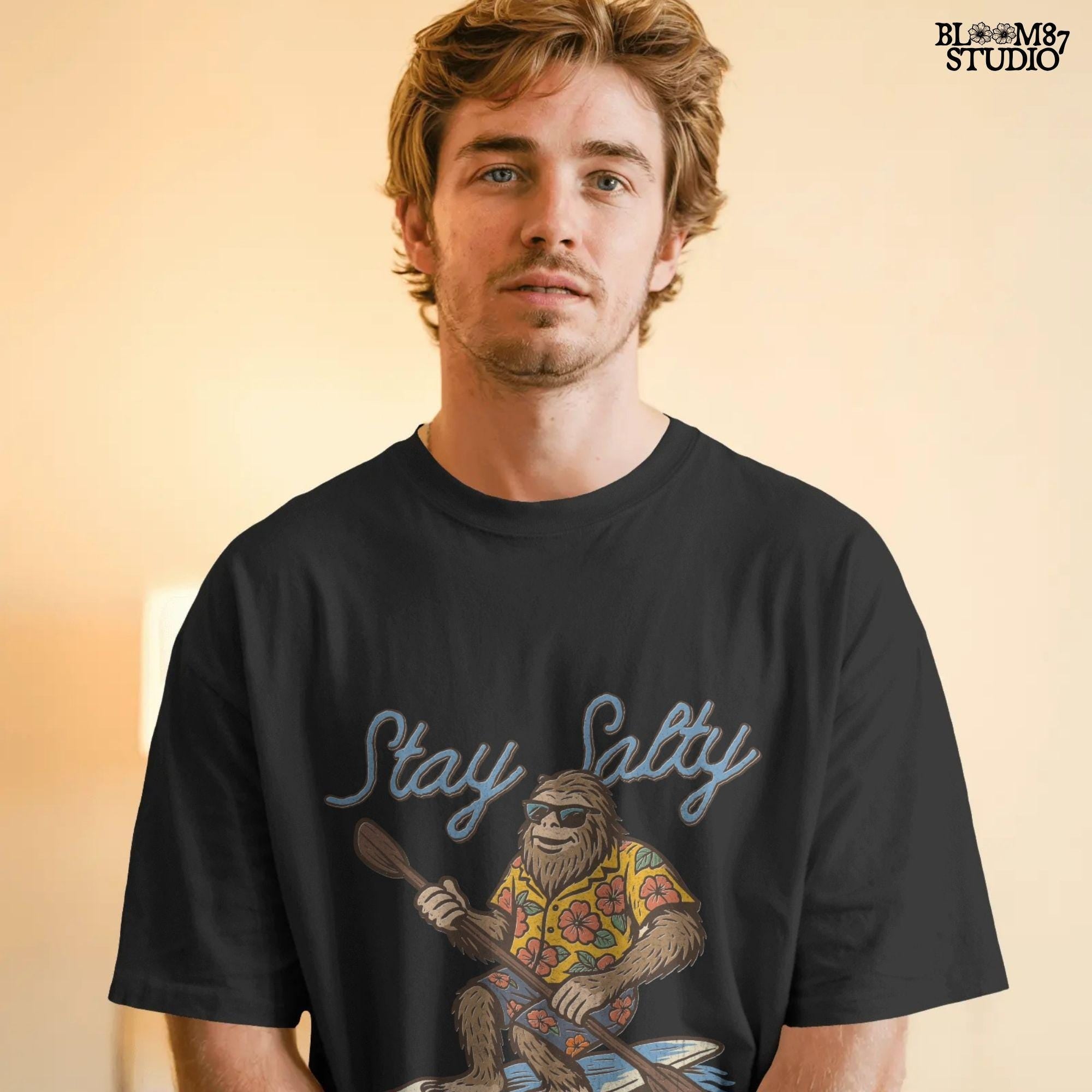 Cartoon Bigfoot wearing a Hawaiian shirt and sunglasses, paddleboarding on a surfboard with text “Stay Salty” – retro beach design PNG for sublimation.