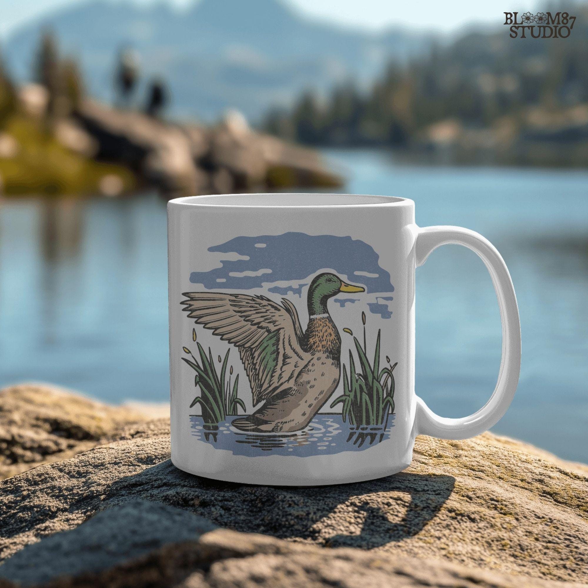 Vintage illustration of a mallard duck with wings spread standing in a pond with reeds and a cloudy blue sky – rustic nature sublimation PNG.