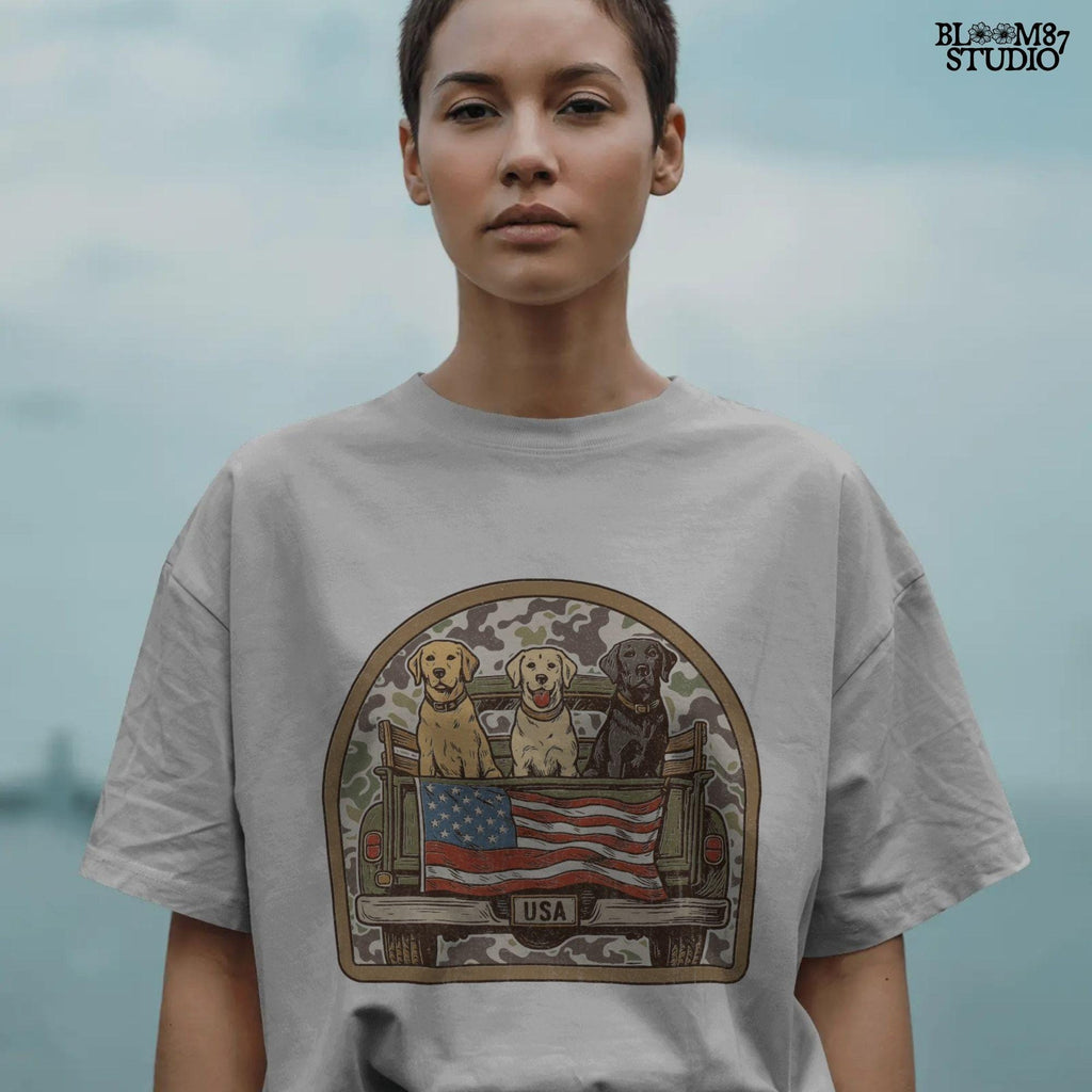 Three Labrador retrievers — yellow, chocolate, and black — sitting on the back of a green camouflage pickup truck with an American flag and the words “Local Boy” underneath.