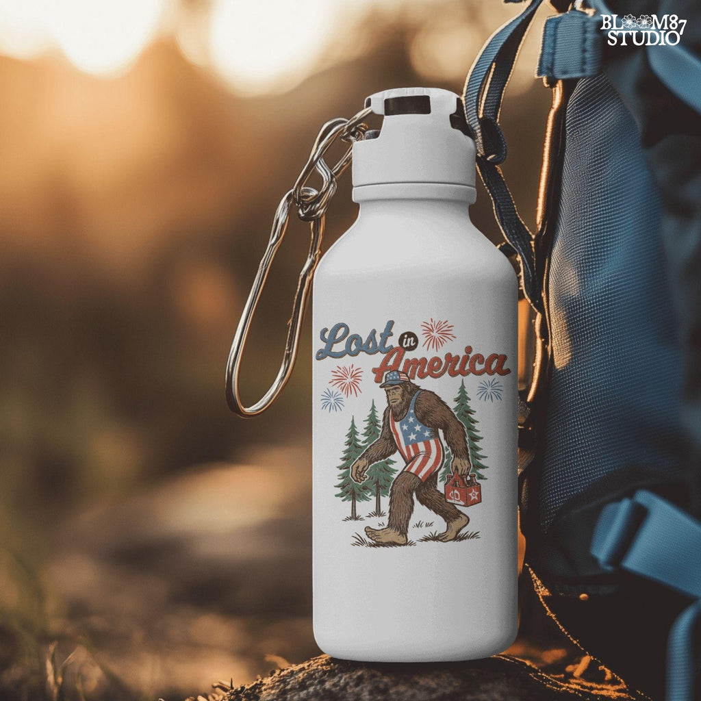 Bigfoot wearing an American flag outfit, carrying a six-pack of beer, walking through pine trees with fireworks in the sky and the phrase “Lost in America.”