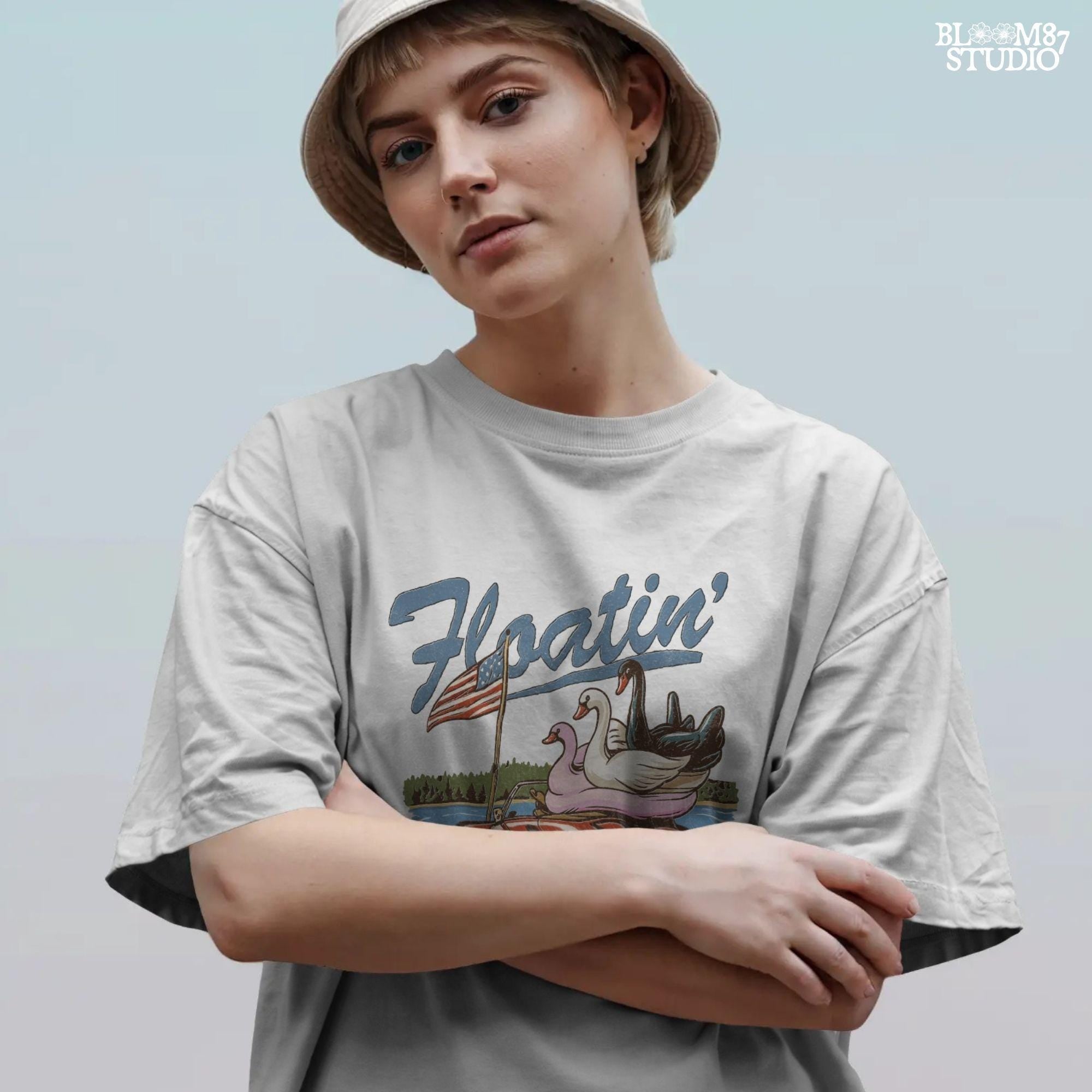 A red convertible car on the beach carrying three swan pool floats with an American flag, with the text “Floatin’ into Freedom” in a playful patriotic theme.