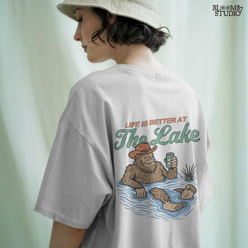 Cartoon Bigfoot relaxing in a lake wearing a cowboy hat, holding a beer can, with retro text saying “Life is Better at the Lake”