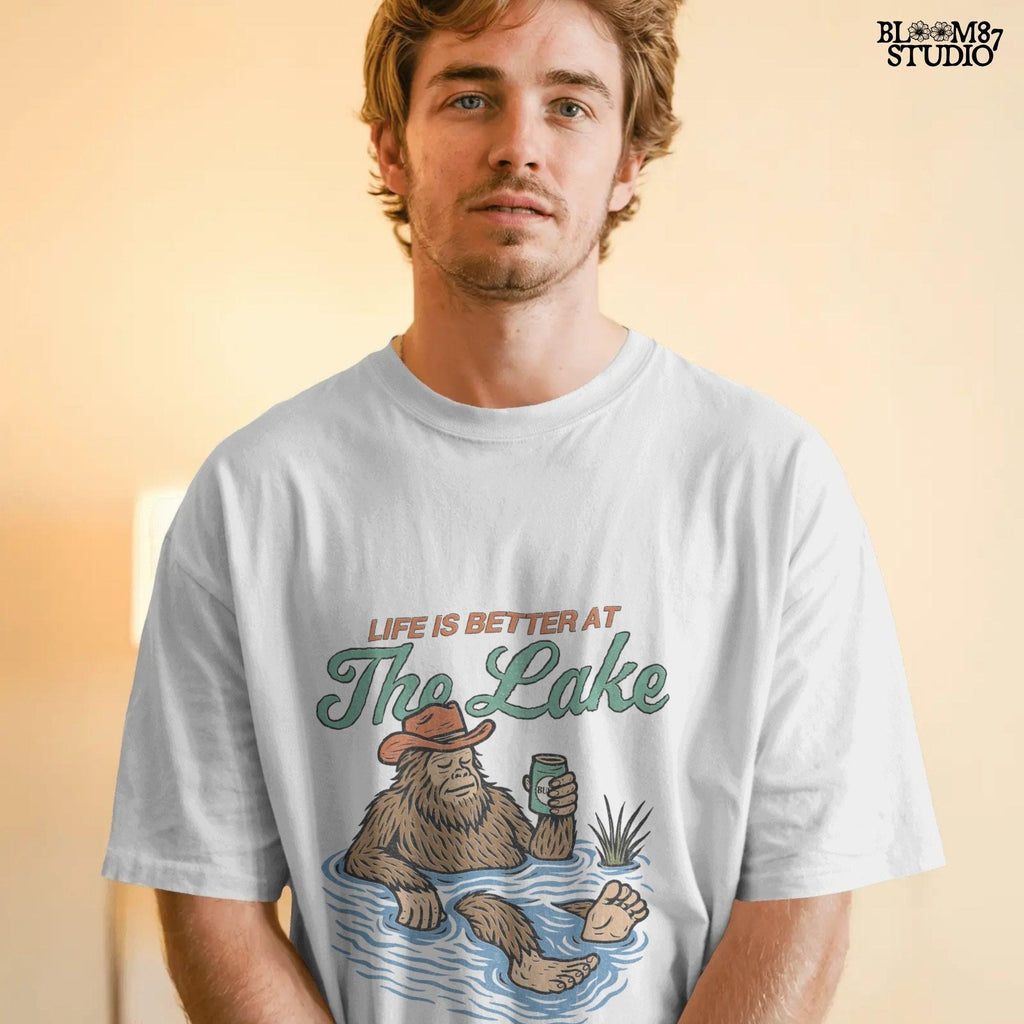 Cartoon Bigfoot relaxing in a lake wearing a cowboy hat, holding a beer can, with retro text saying “Life is Better at the Lake”