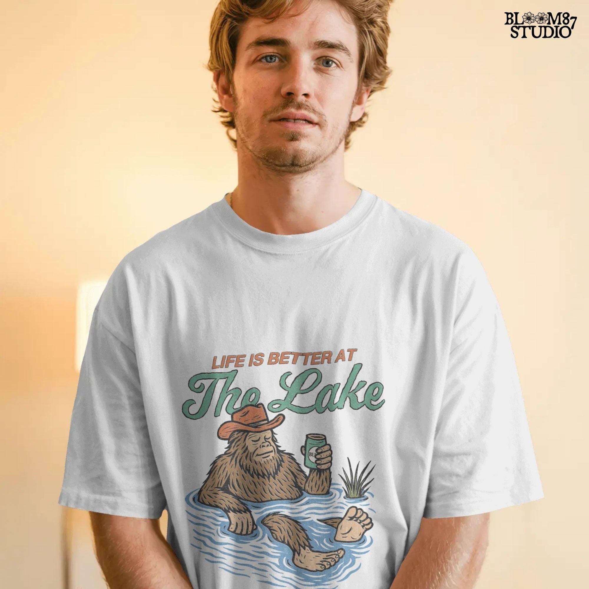 Cartoon Bigfoot relaxing in a lake wearing a cowboy hat, holding a beer can, with retro text saying “Life is Better at the Lake”