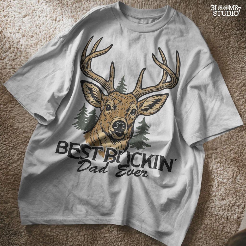 Rustic illustration of a buck deer with antlers and pine trees, text below says “Best Buckin’ Dad Ever” in bold and script font