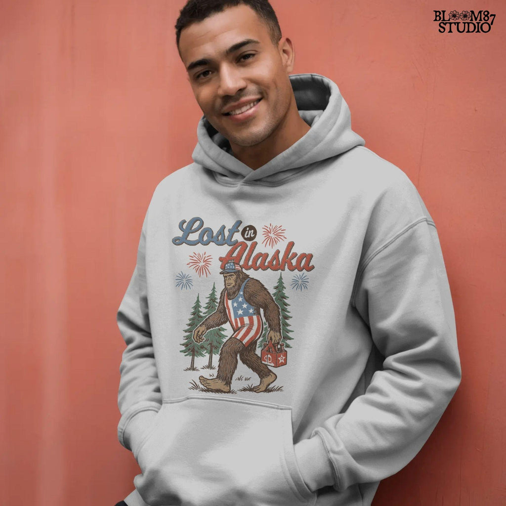 Bigfoot wearing an American flag outfit and cap, walking through forest with fireworks in background, holding a six-pack of beer with “Lost in Alaska” text