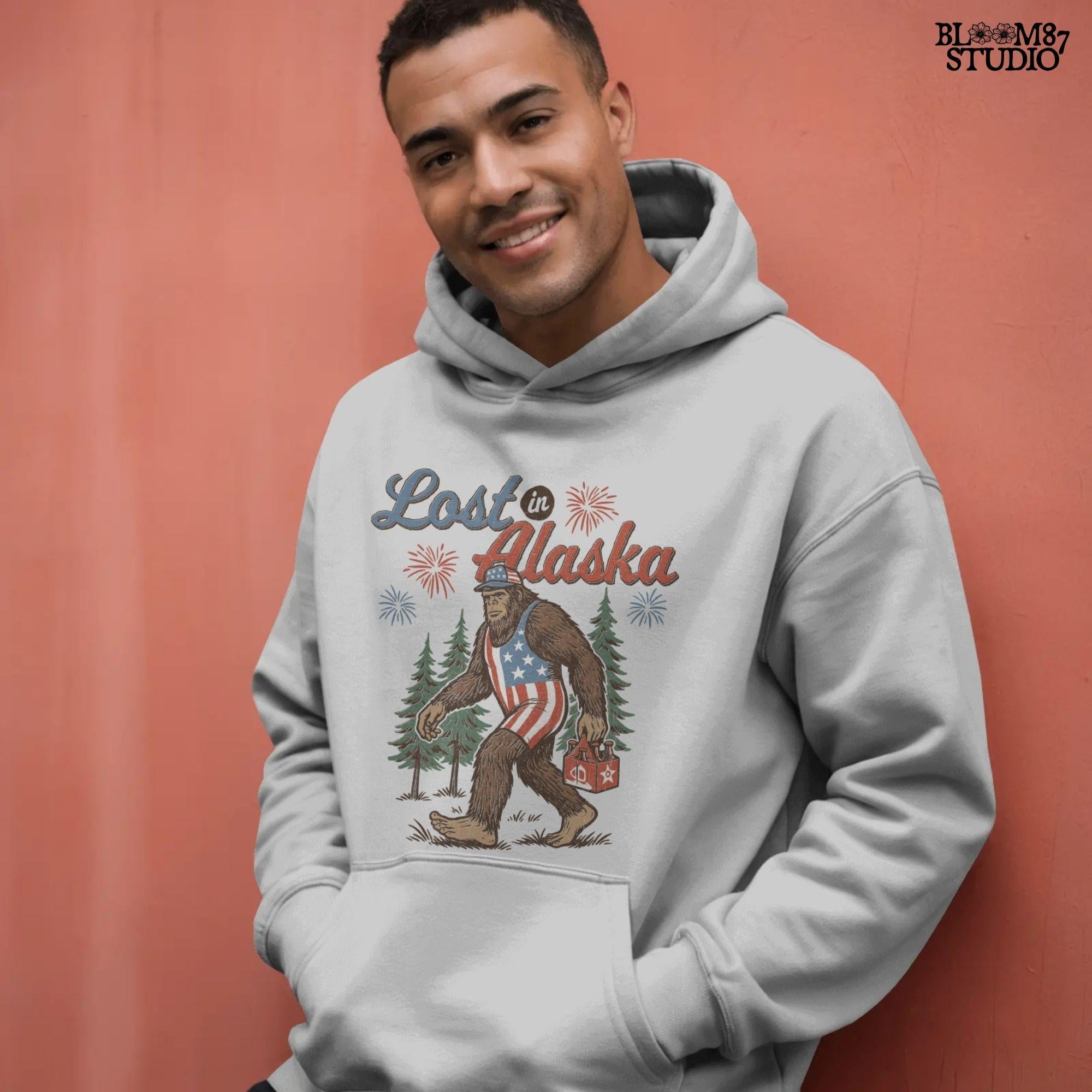 Bigfoot wearing an American flag outfit and cap, walking through forest with fireworks in background, holding a six-pack of beer with “Lost in Alaska” text