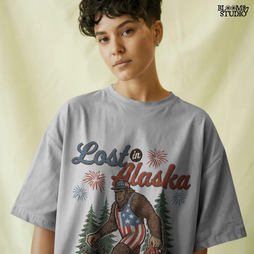 Bigfoot wearing an American flag outfit and cap, walking through forest with fireworks in background, holding a six-pack of beer with “Lost in Alaska” text