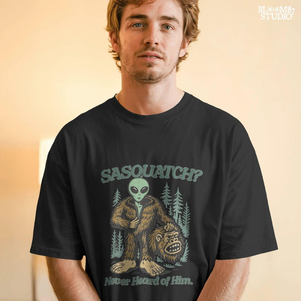Funny alien unzipping a Bigfoot costume with Sasquatch head in hand, forest trees in background, and text “Sasquatch? Never Heard of Him.”