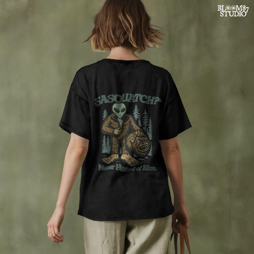Funny alien unzipping a Bigfoot costume with Sasquatch head in hand, forest trees in background, and text “Sasquatch? Never Heard of Him.”